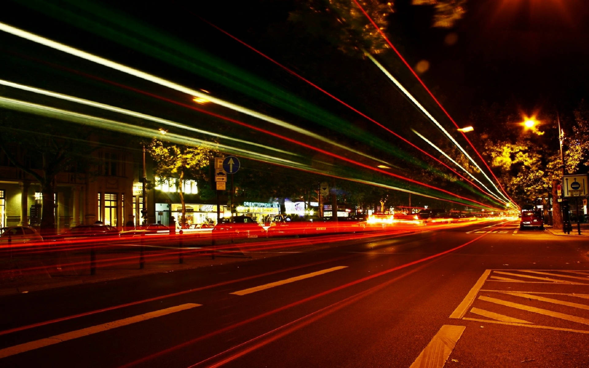柏林城市道路 壁紙 — 紅色、綠色和白色的鮮豔光帶在夜間濕漉漉的城市街道上模糊地掠過，捕捉到發光的商店櫥窗和樹木前的動感。
