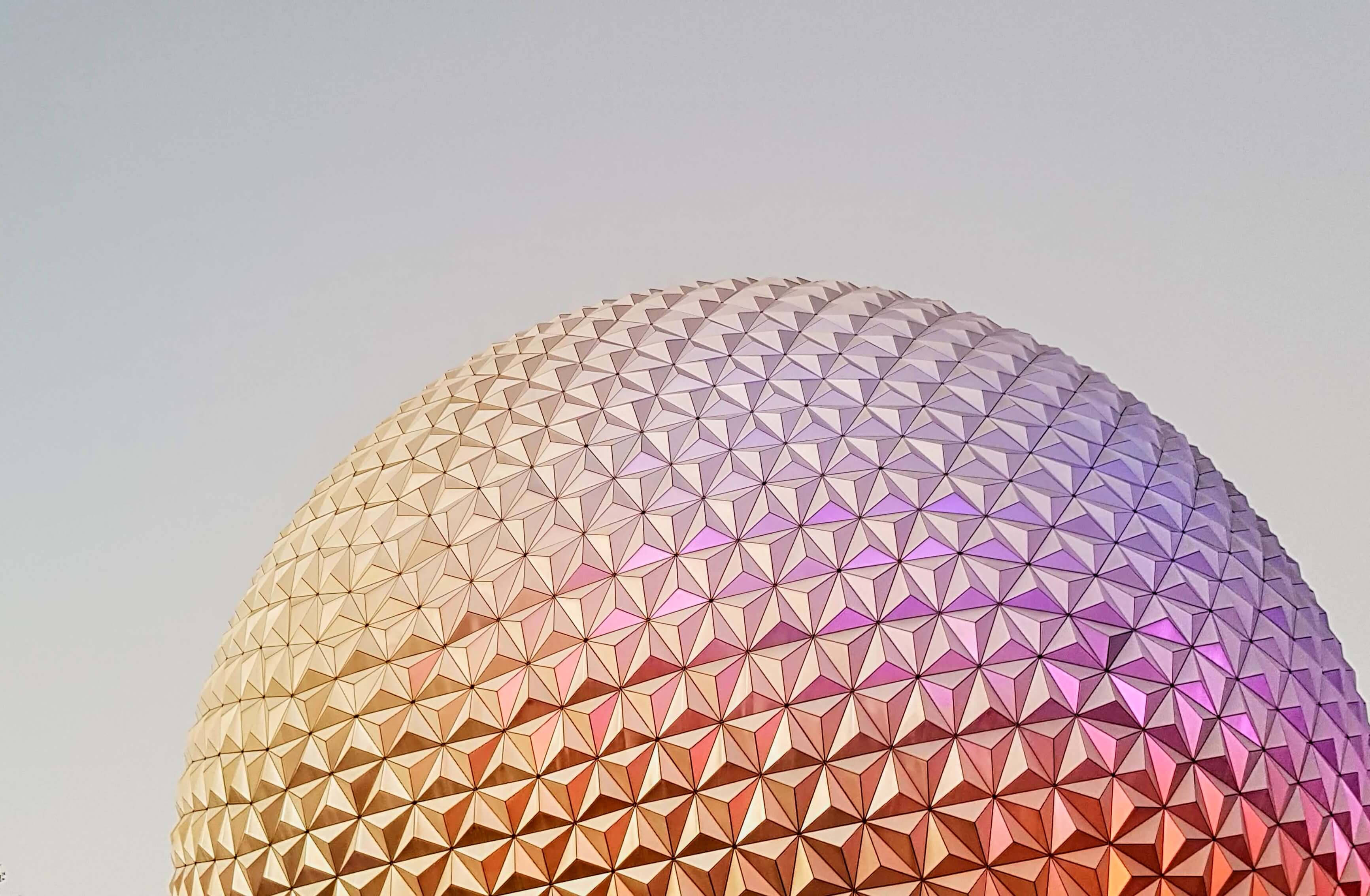 柔和漸層的Epcot球體 壁紙 — 一個測地線穹頂的特寫，其三角形面在淡色天空的映襯下散發出溫暖的橙色和冷紫色調的光芒。