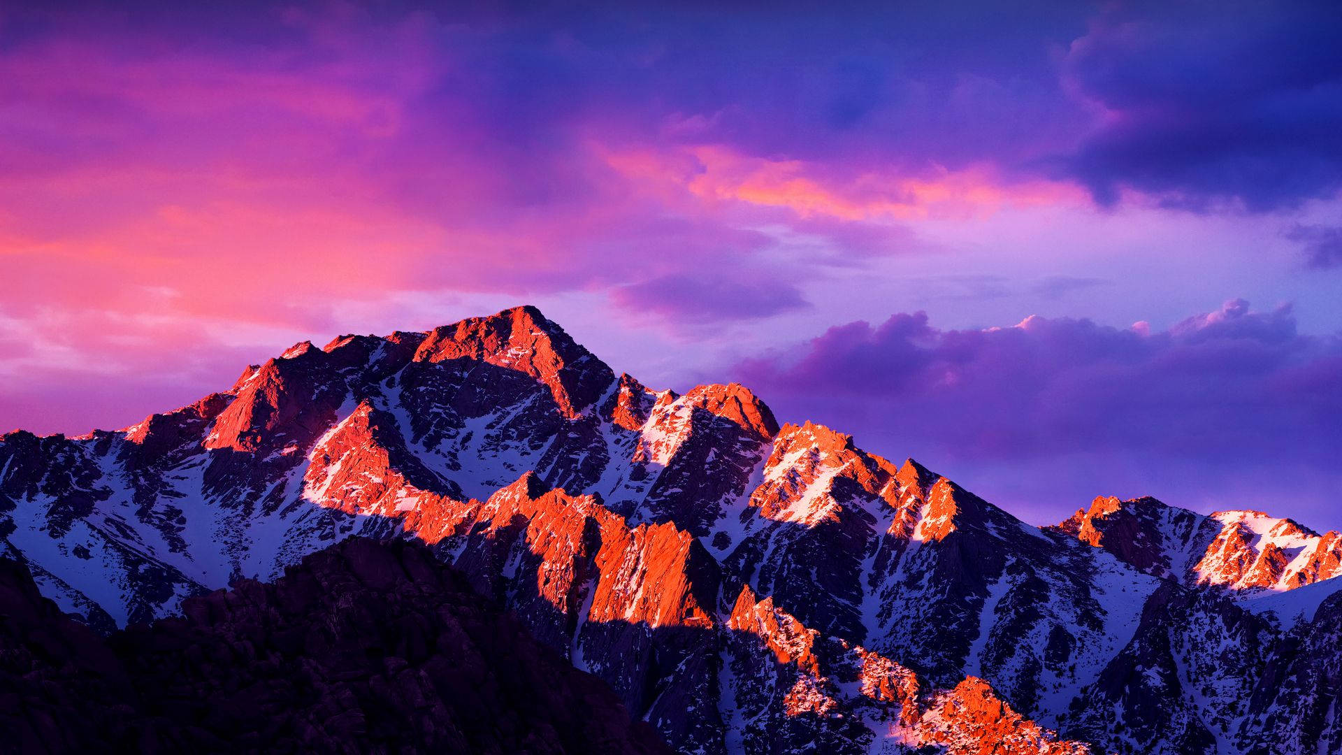 柔粉山景 macOS 壁紙 — 白雪覆蓋的山峰在戲劇性的紫色和粉紅色日落天空映襯下，閃耀著火橙色的光芒。