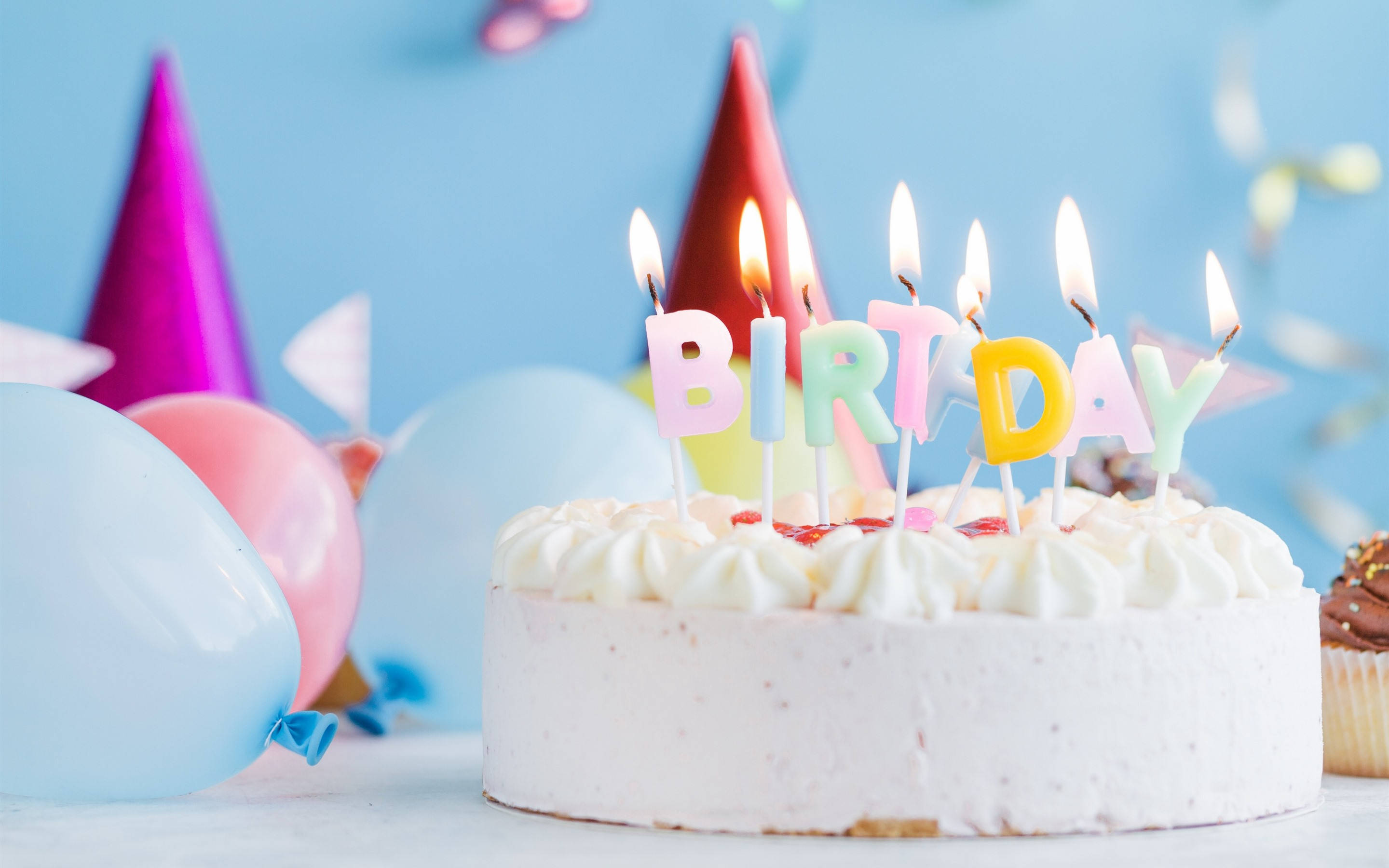 柔美白色生日蛋糕與點燃蠟燭 壁紙 — 一個糖霜生日蛋糕，頂部插著彩色「BIRTHDAY」蠟燭，周圍環繞著派對氣球，背景是柔和的藍色。