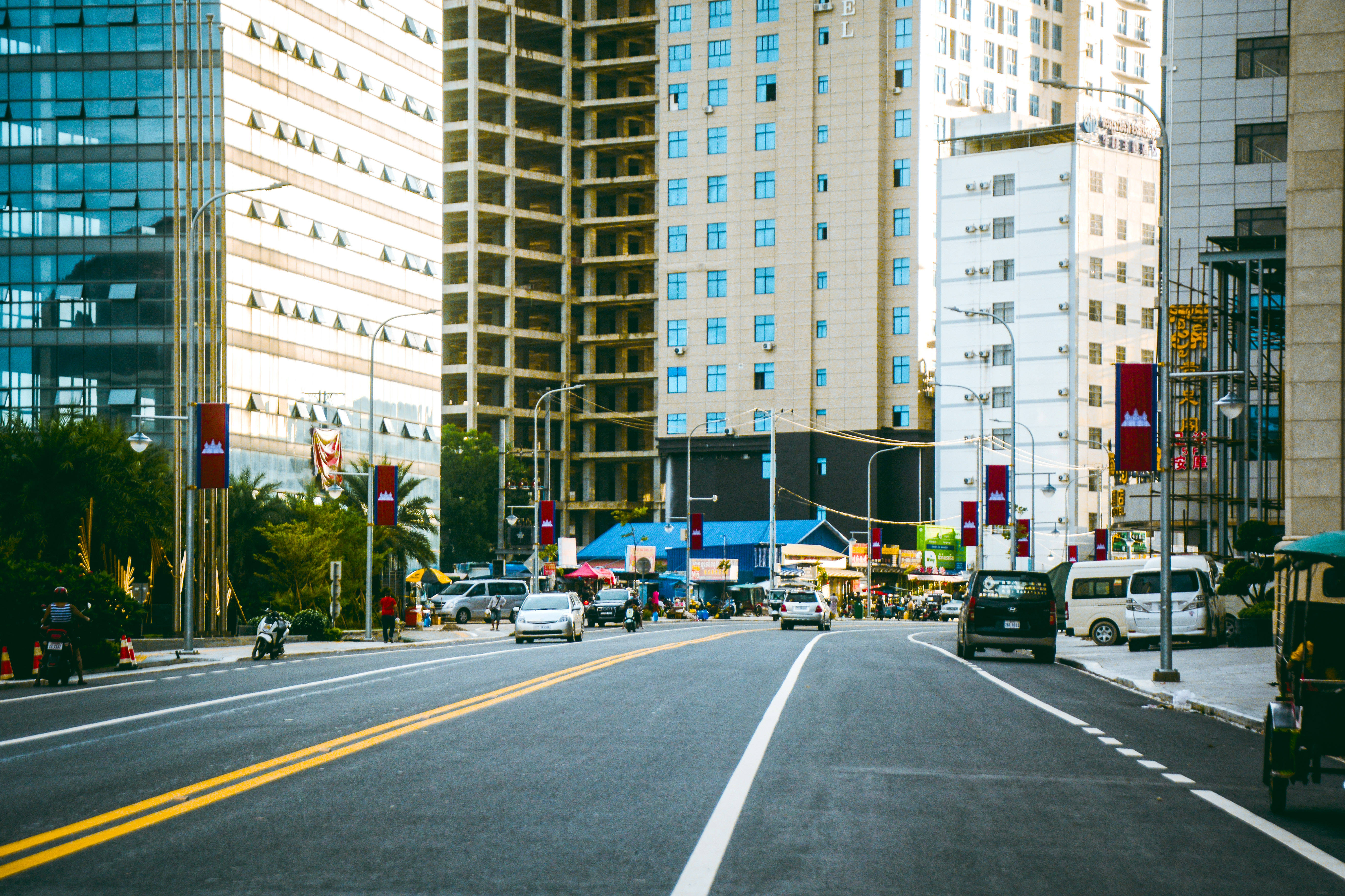 柬埔寨高樓大廈 壁紙 — 一條寬闊的城市街道，兩旁是現代化的玻璃和混凝土摩天大樓，藍色遮陽篷下是熙熙攘攘的路邊攤販。
