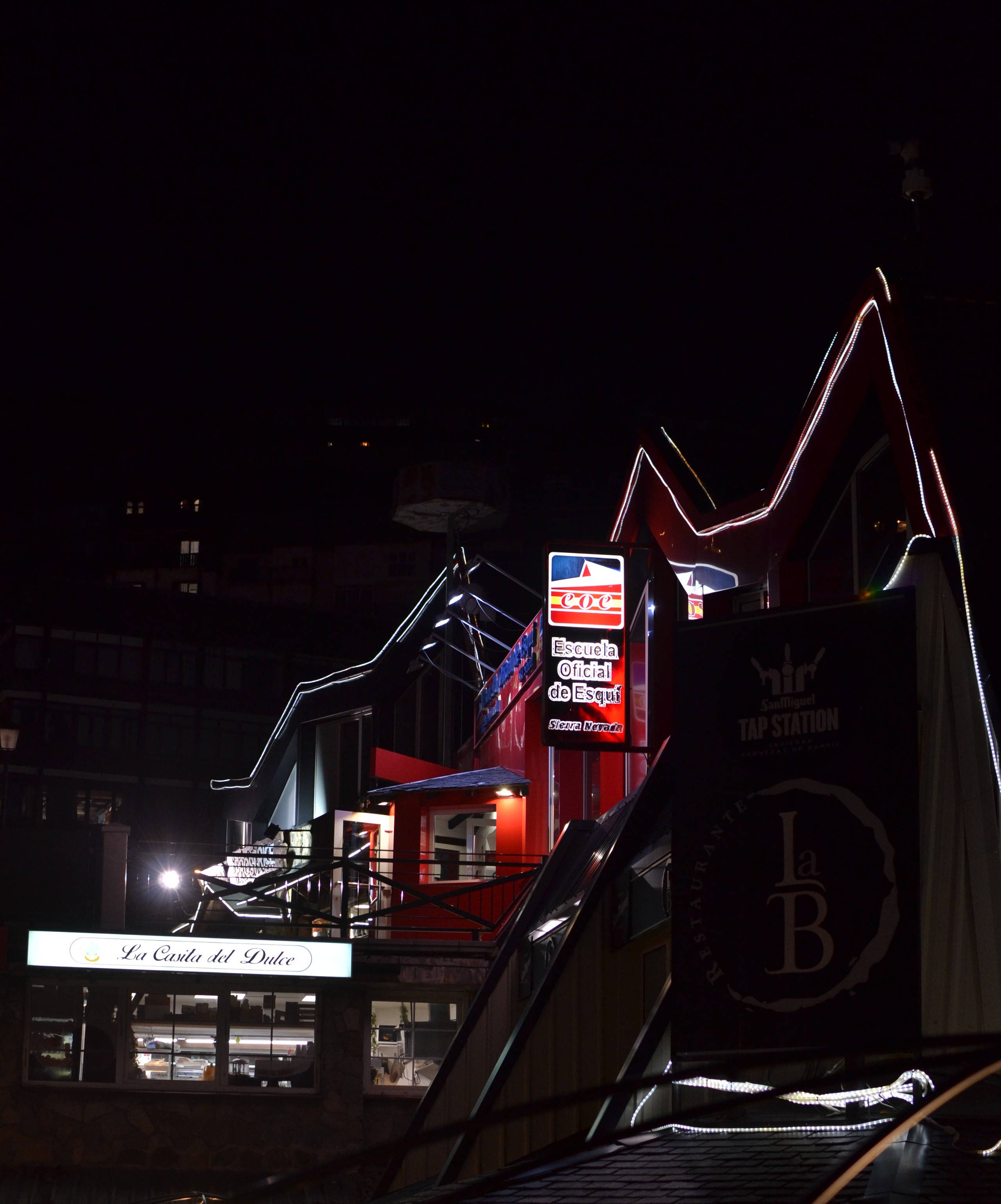 格林納達夜景城市 壁紙 — 一個充滿活力的夜景，照亮了滑雪勝地建築物，帶有發光的標誌和戲劇性的紅色燈光，映襯著黑暗的天空。
