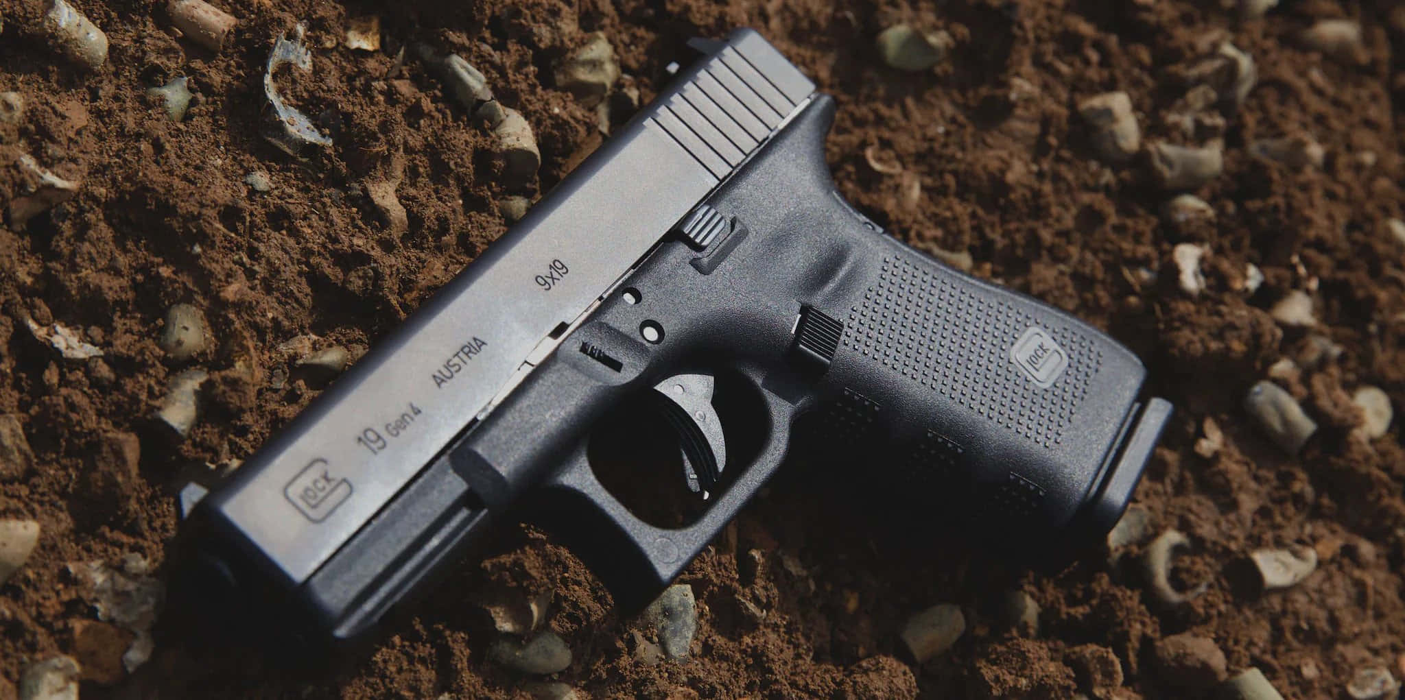 格洛克19手槍的高解析度特寫圖像 壁紙 — 一把黑色Glock 19 Gen4手槍在明亮日光下放置於粗糙的棕色泥土上，散落著小石子。