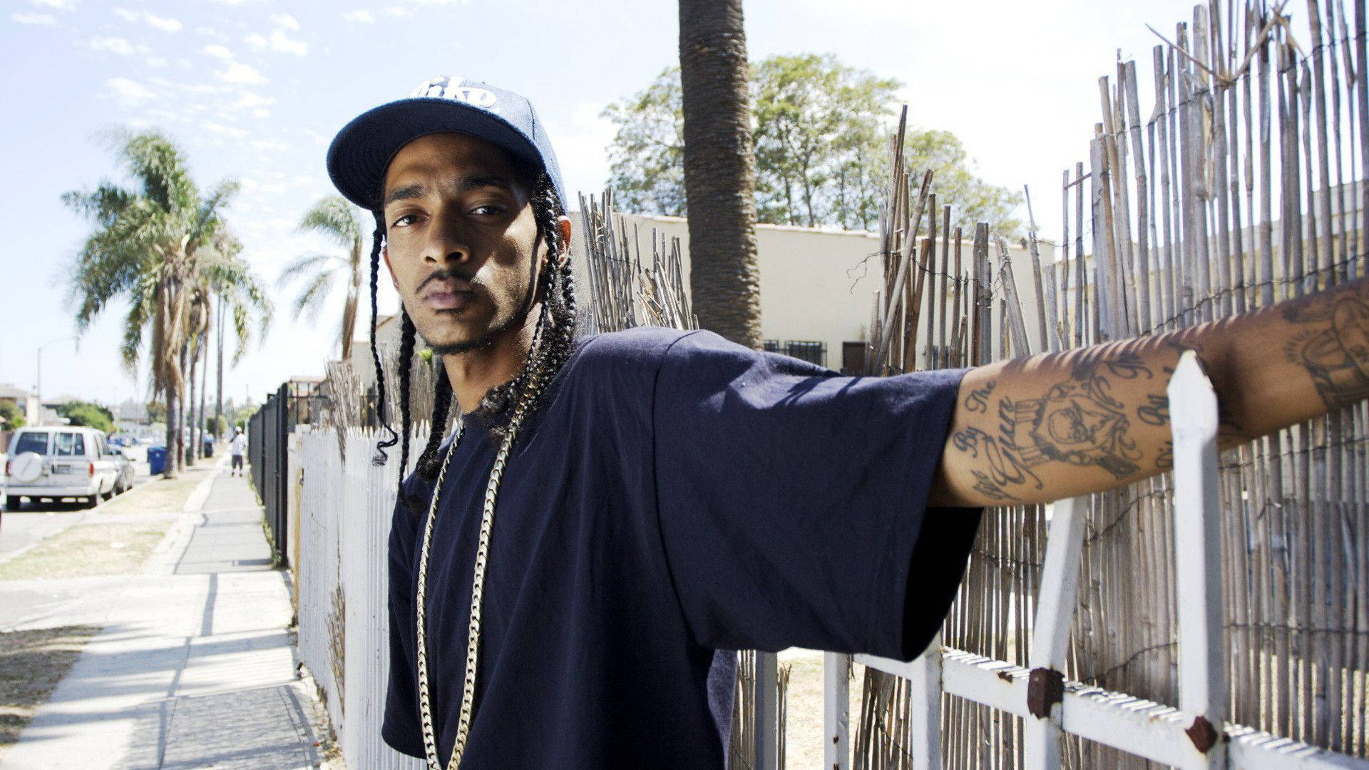 格萊美得主饒舌歌手 Nipsey Hussle 演唱會現場 壁紙 — 一個梳辮子、有紋身的男人，在陽光明媚、棕櫚樹成蔭的街道上靠著白色柵欄。