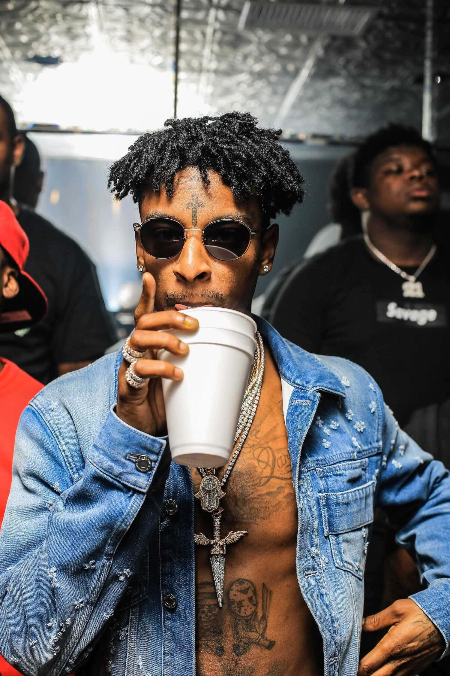 格萊美獎得主饒舌歌手 21 Savage 壁紙 — 一位時尚男士，留著辮子頭和紋身，戴著太陽眼鏡和牛仔外套，正從白色杯子中啜飲。