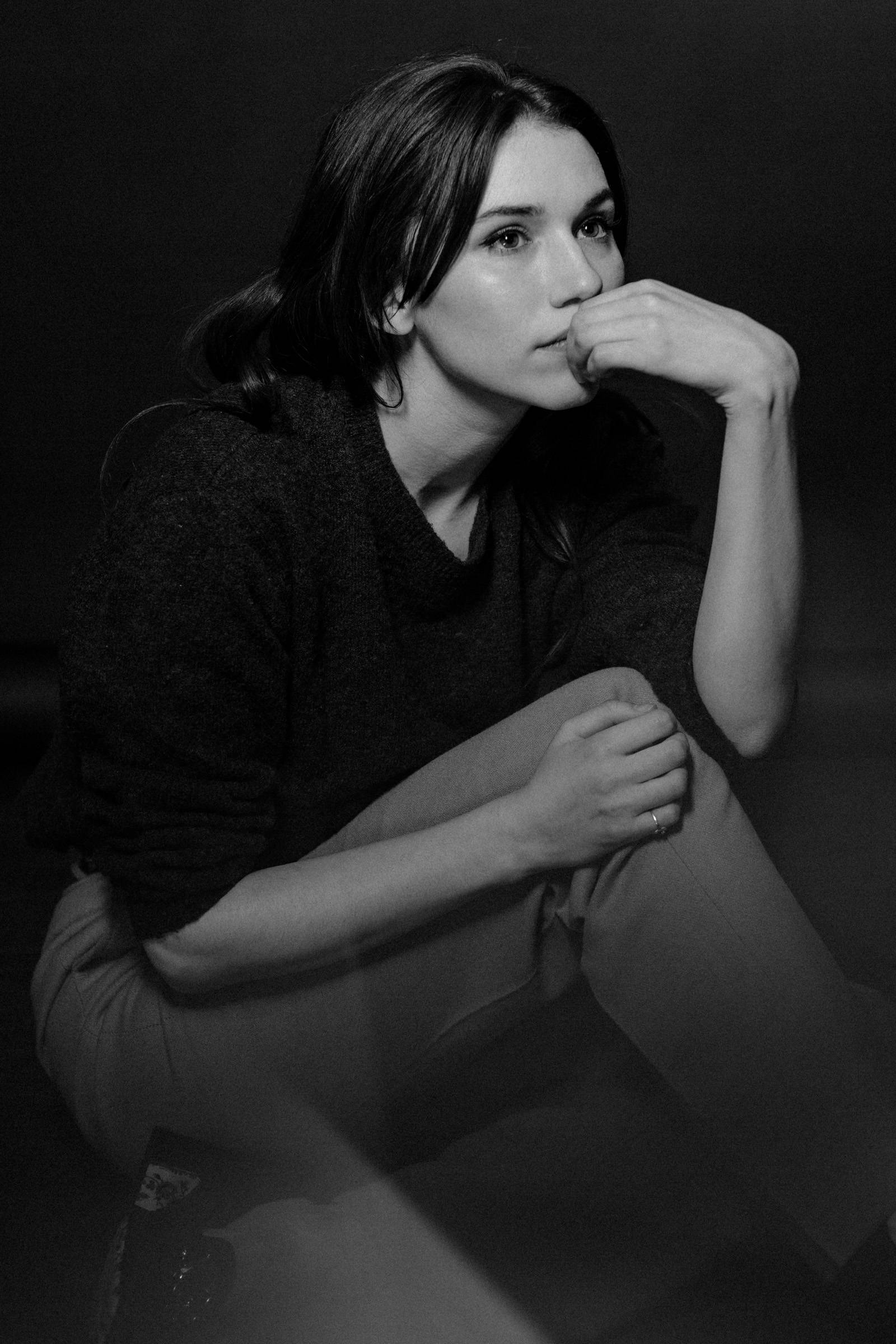 格蕾絲·富爾頓黑白肖像 壁紙 — 一位沉思的黑白女子坐著，手靠近嘴巴，在深色背景下若有所思地凝視。