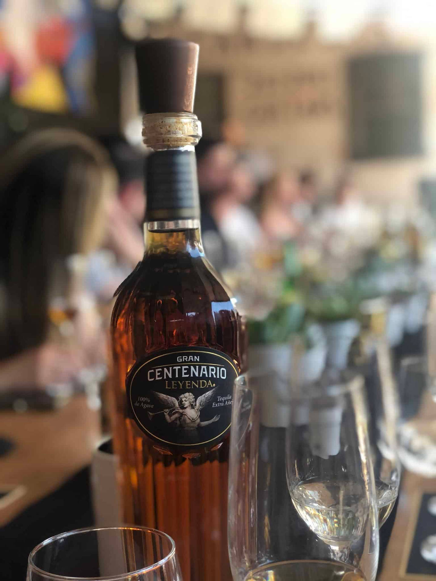 一瓶 Gran Centenario Leyenda 龍舌蘭酒放在酒吧上，背景有空杯子和模糊的顧客。
