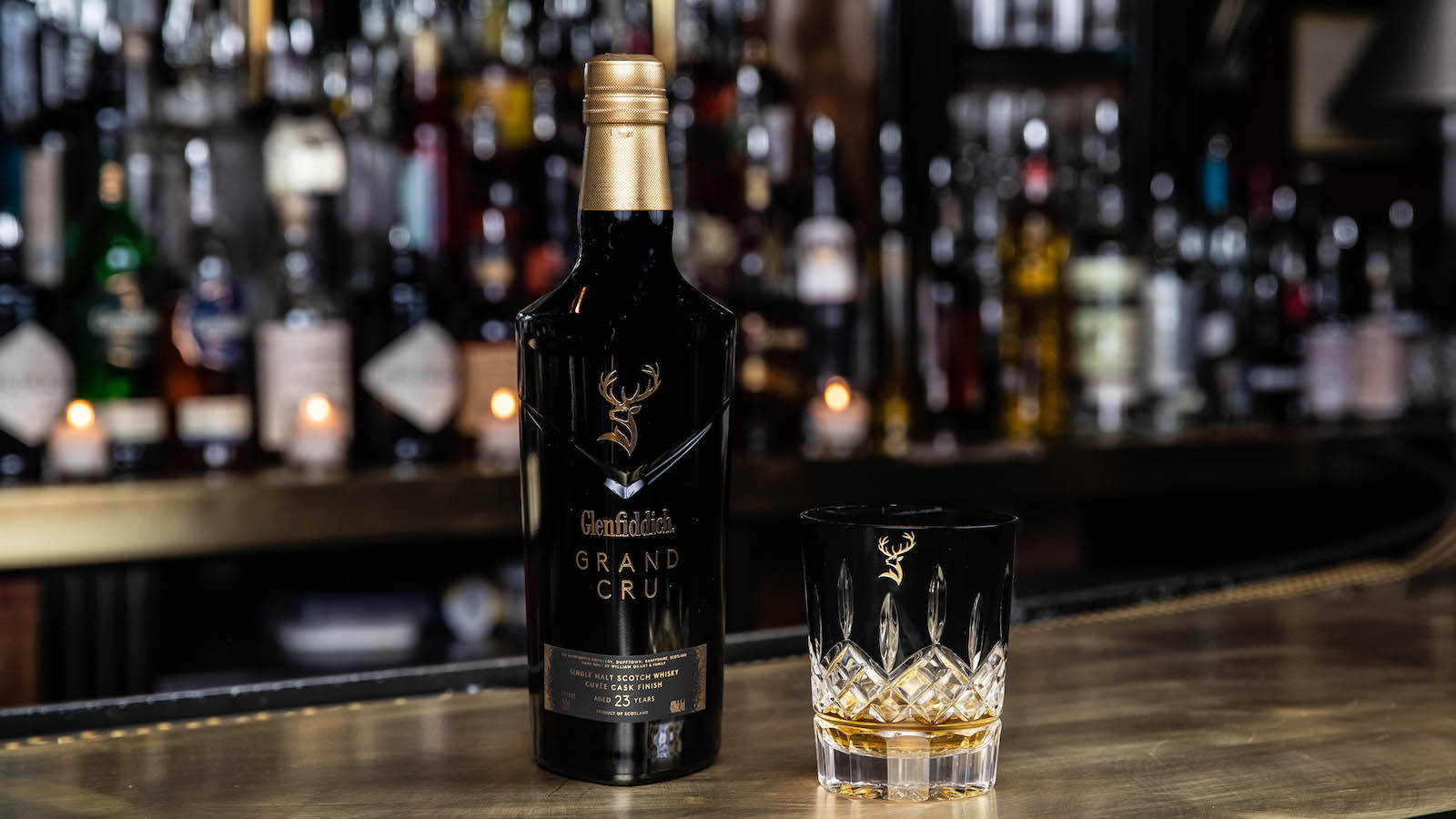 酒吧上的 Glenfiddich Grand Cru 威士忌酒瓶和酒杯，後面有蠟燭和酒瓶。