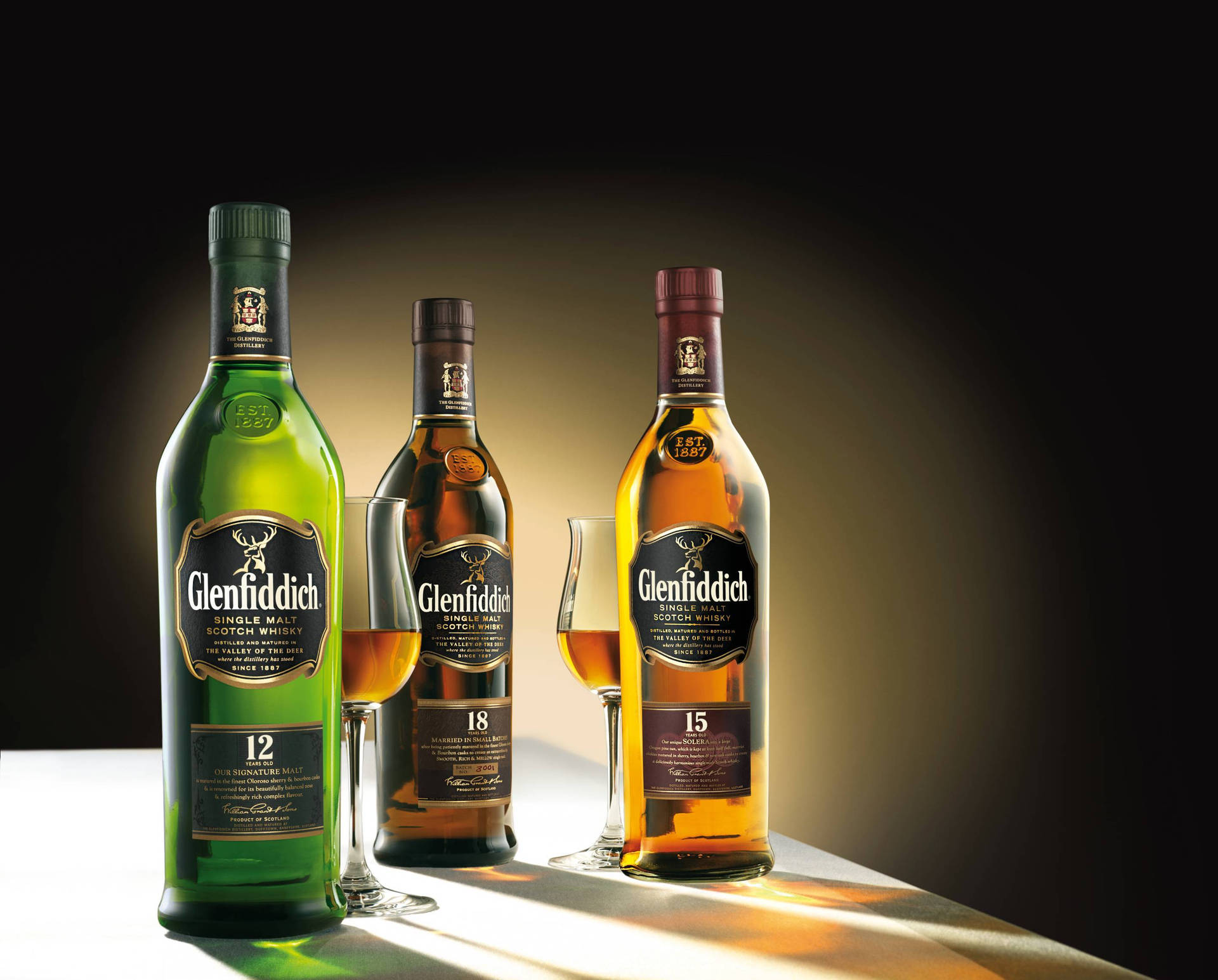 三瓶 Glenfiddich 威士忌酒瓶連酒杯放在發光表面上，背景黑暗。