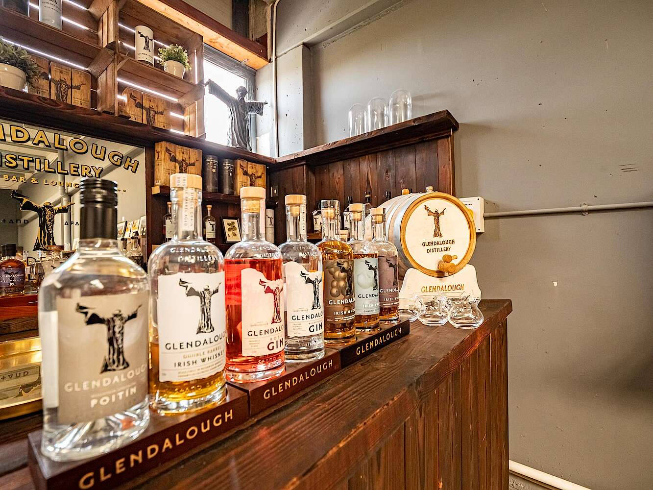 格蘭達洛野生玫瑰金酒於酒吧場景 壁紙 — 一個木製吧枱展示Glendalough烈酒瓶，配有品牌酒杯和陳列商品的貨架，位於質樸的釀酒廠休息室。