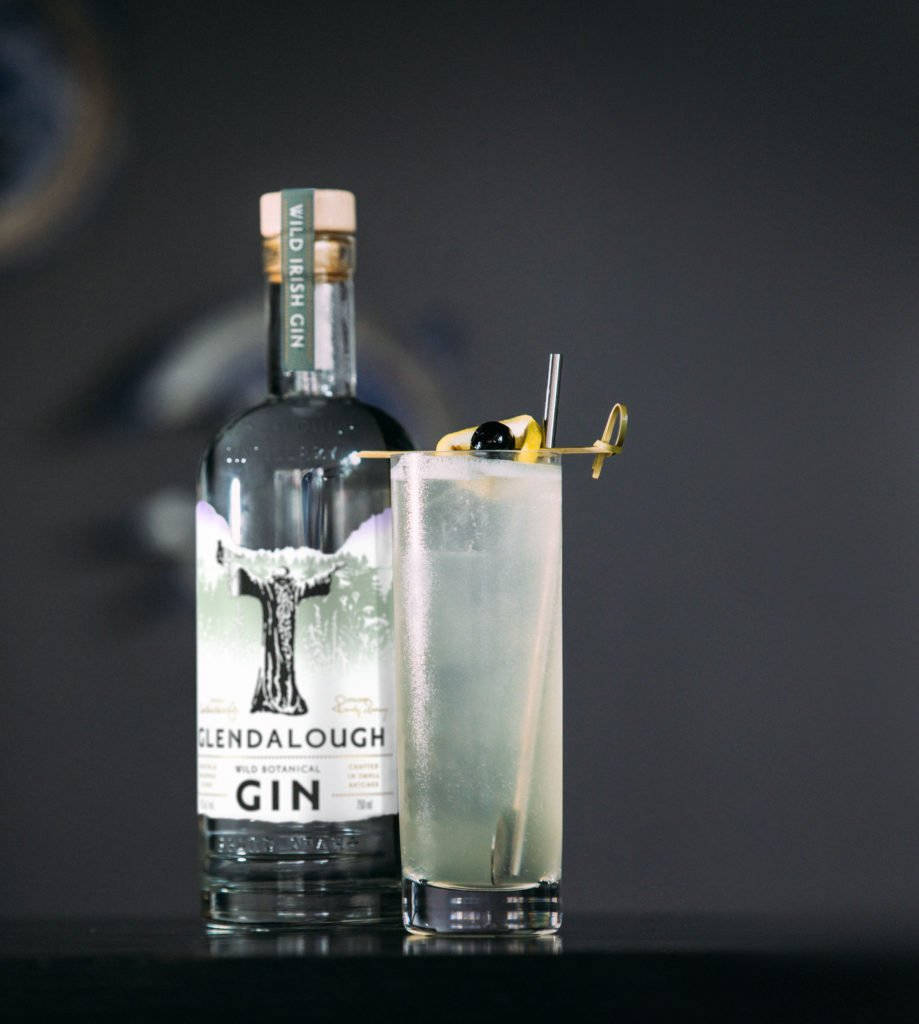 格蘭達洛野生琴酒湯姆·柯林斯雞尾酒 壁紙 — 一瓶時尚的Glendalough Wild Irish Gin旁邊放著一杯高身氈酒雞尾酒，飾以檸檬扭紋和橄欖。