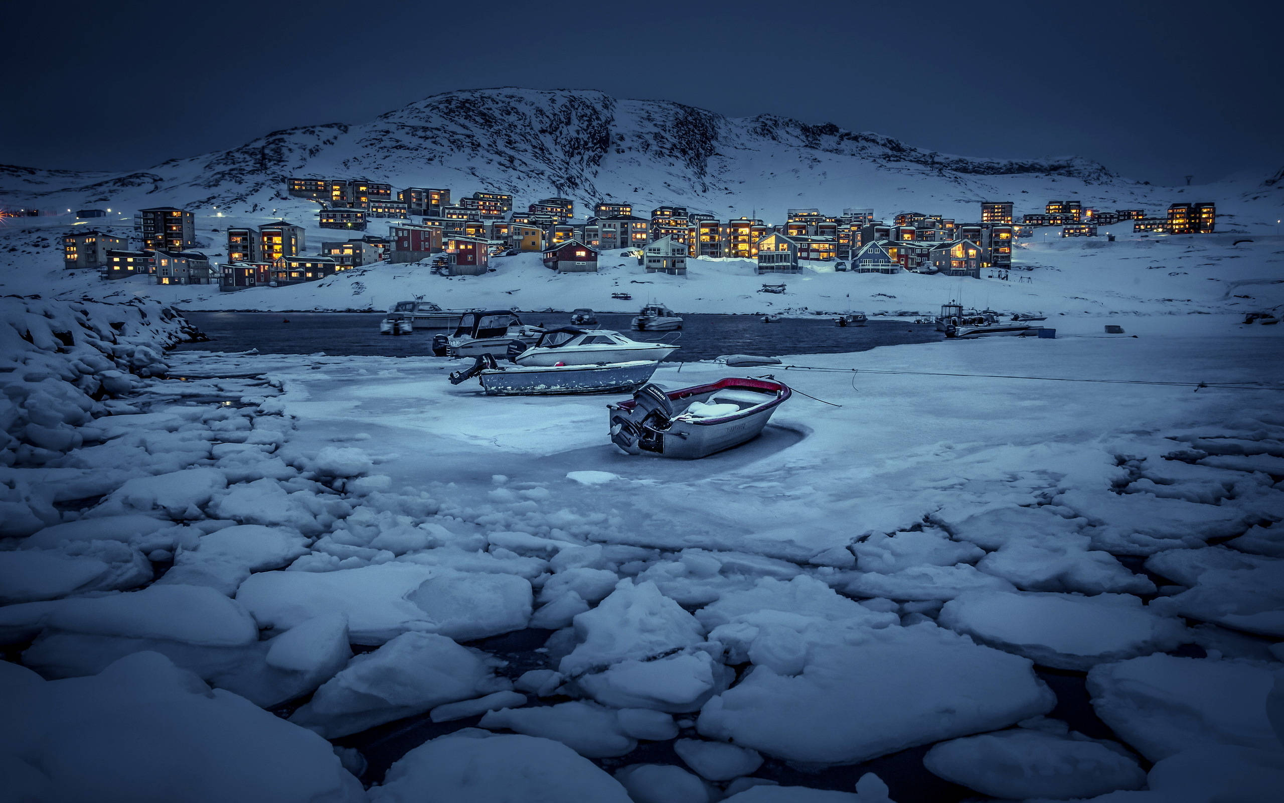 格陵蘭夜間風景 壁紙 — 被雪覆蓋的船隻停泊在冰冷的水面上，溫暖的燈光從依附在雪坡上的房屋中在黃昏時分閃爍。