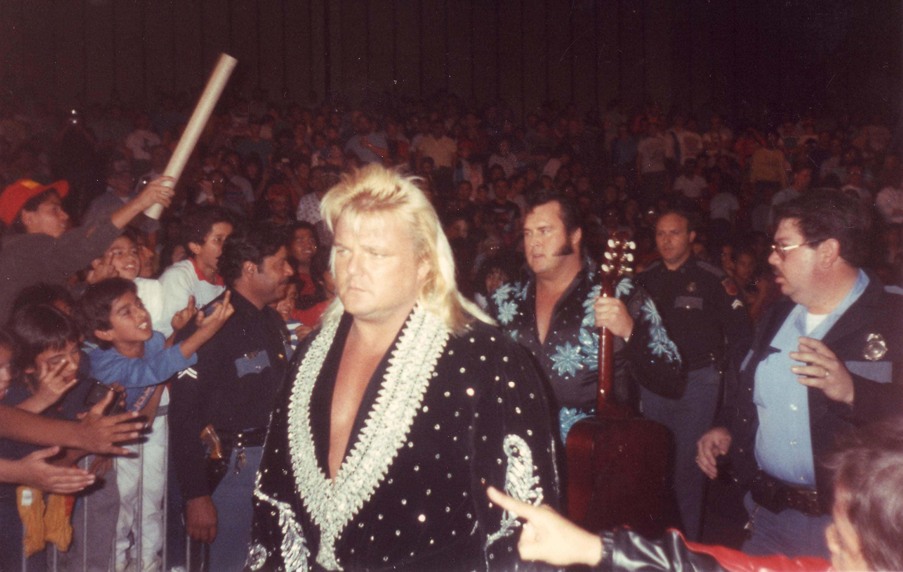 格雷格·瓦倫丁與Honky Tonk Man 壁紙 — 一位金髮、身穿閃亮黑色外套的摔角手，在擠滿人的場館中穿過歡呼的人群。