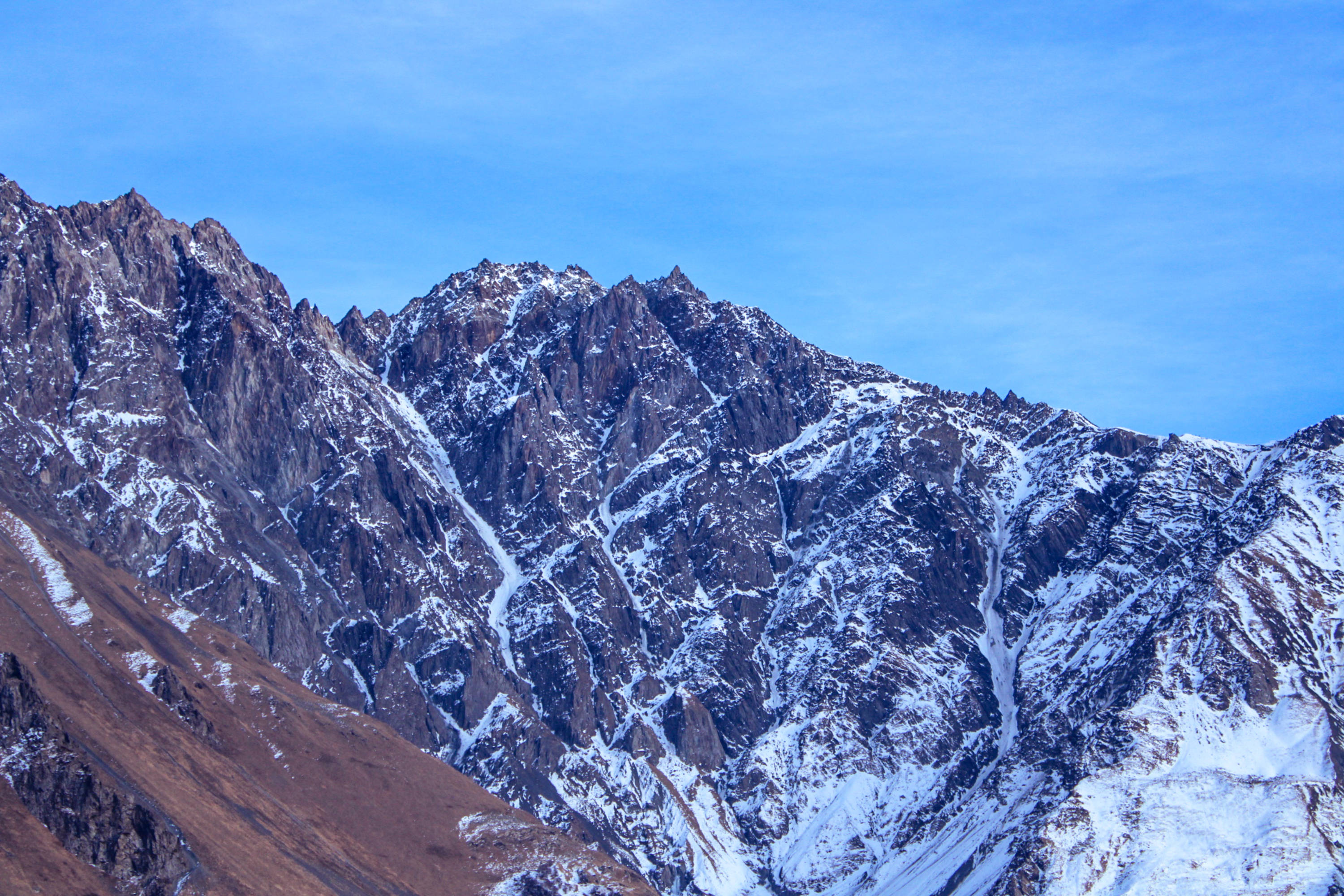 格魯吉亞斯捷潘茨明達山 壁紙 — 鋸齒狀的雪山山脈在清澈的藍天和稀疏的縷縷白雲襯托下巍然聳立。