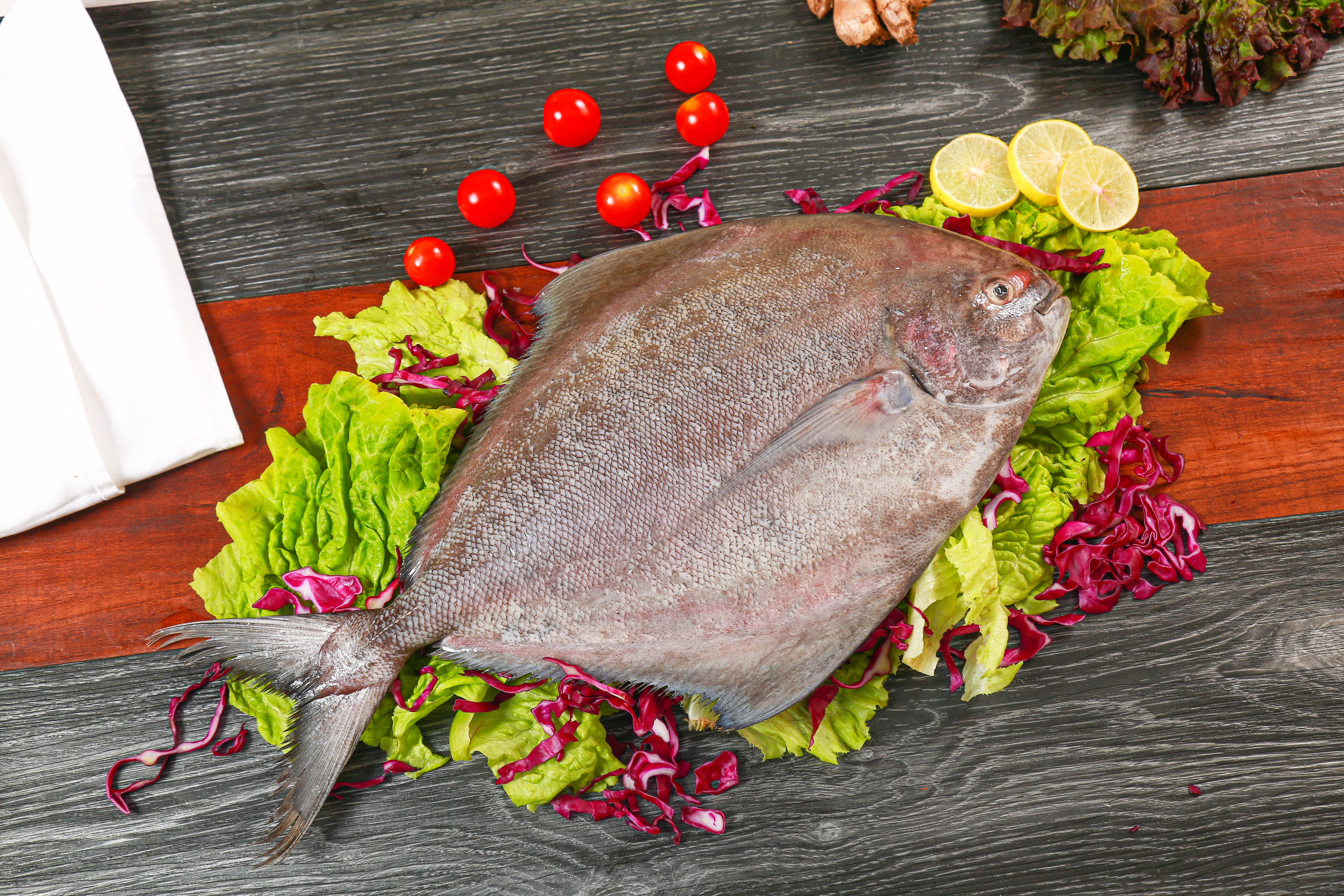 桌上平鋪鮮魚鰈 壁紙 — 一條完整的銀色鰈魚躺在綠色和紫色生菜床上，配上櫻桃番茄和檸檬片，放在深色木質表面上。