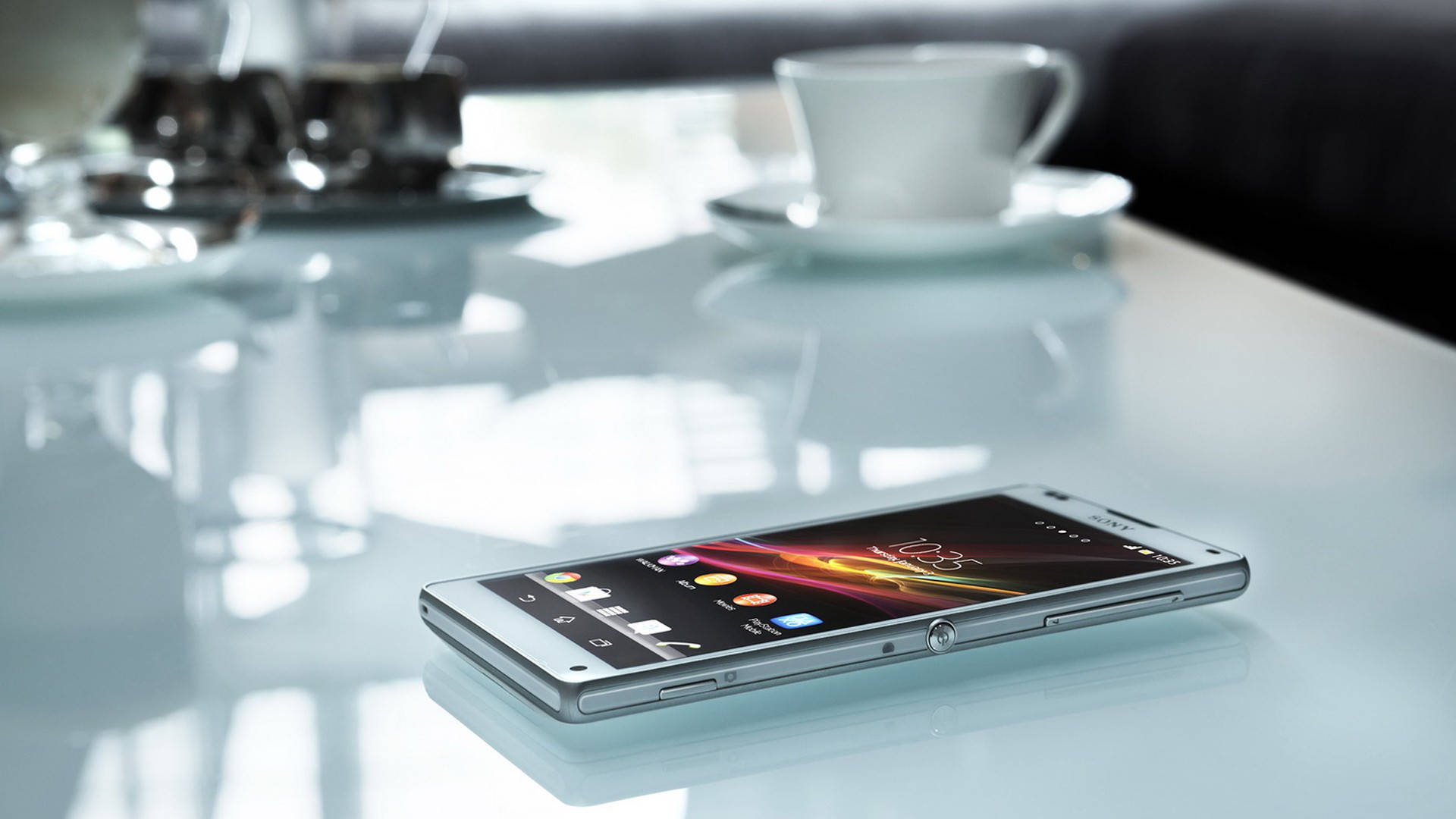 桌上索尼Xperia 壁紙 — 一部時尚的銀色智能手機放在光亮的白色桌子上，旁邊有咖啡杯和玻璃器皿，環境明亮現代。