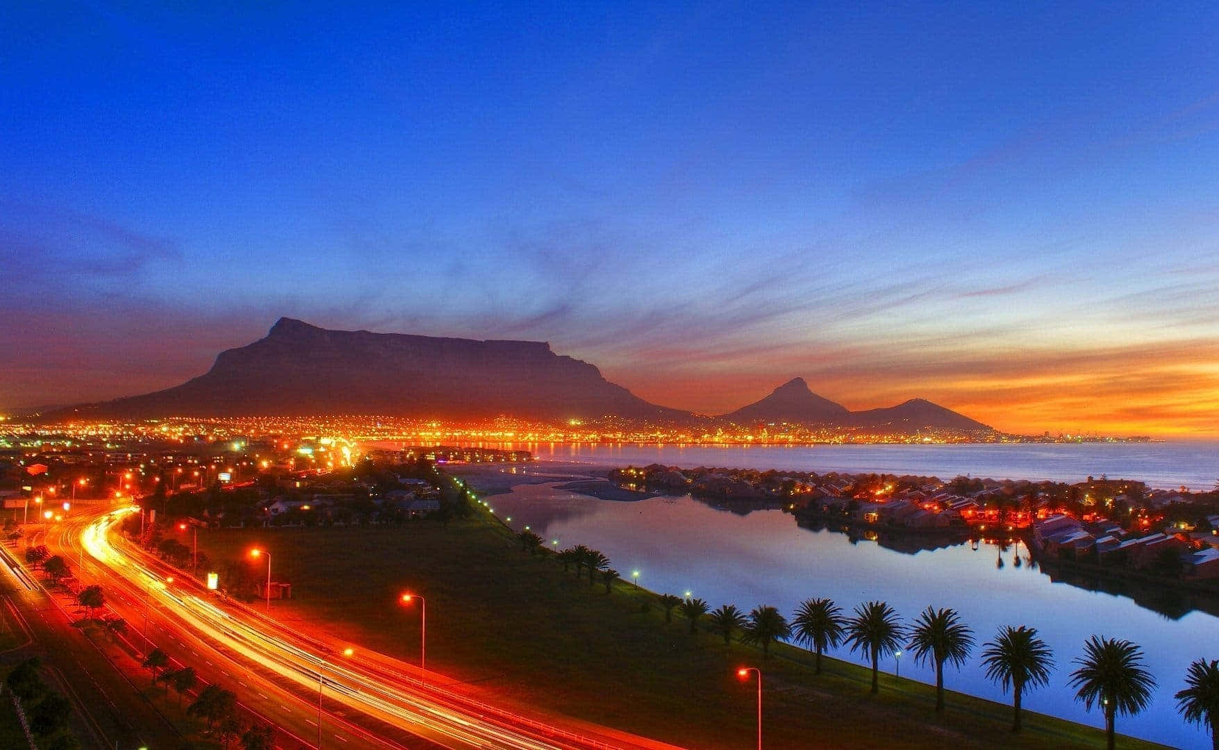 South Africa 桌布 壁紙 — 桌山在黃昏時分聳立在充滿活力的城市天際線上，紅色車燈劃過，平靜的水面反射著溫暖的橙色和藍色天空。