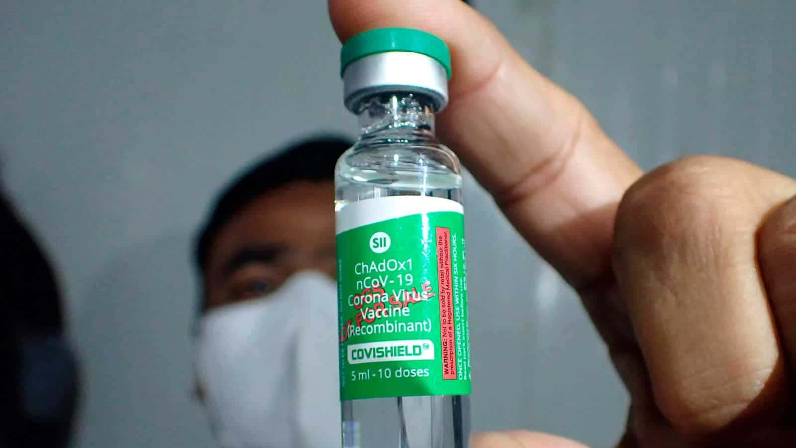 特寫一隻手拿著Covishield疫苗小瓶，背景有戴口罩的人。