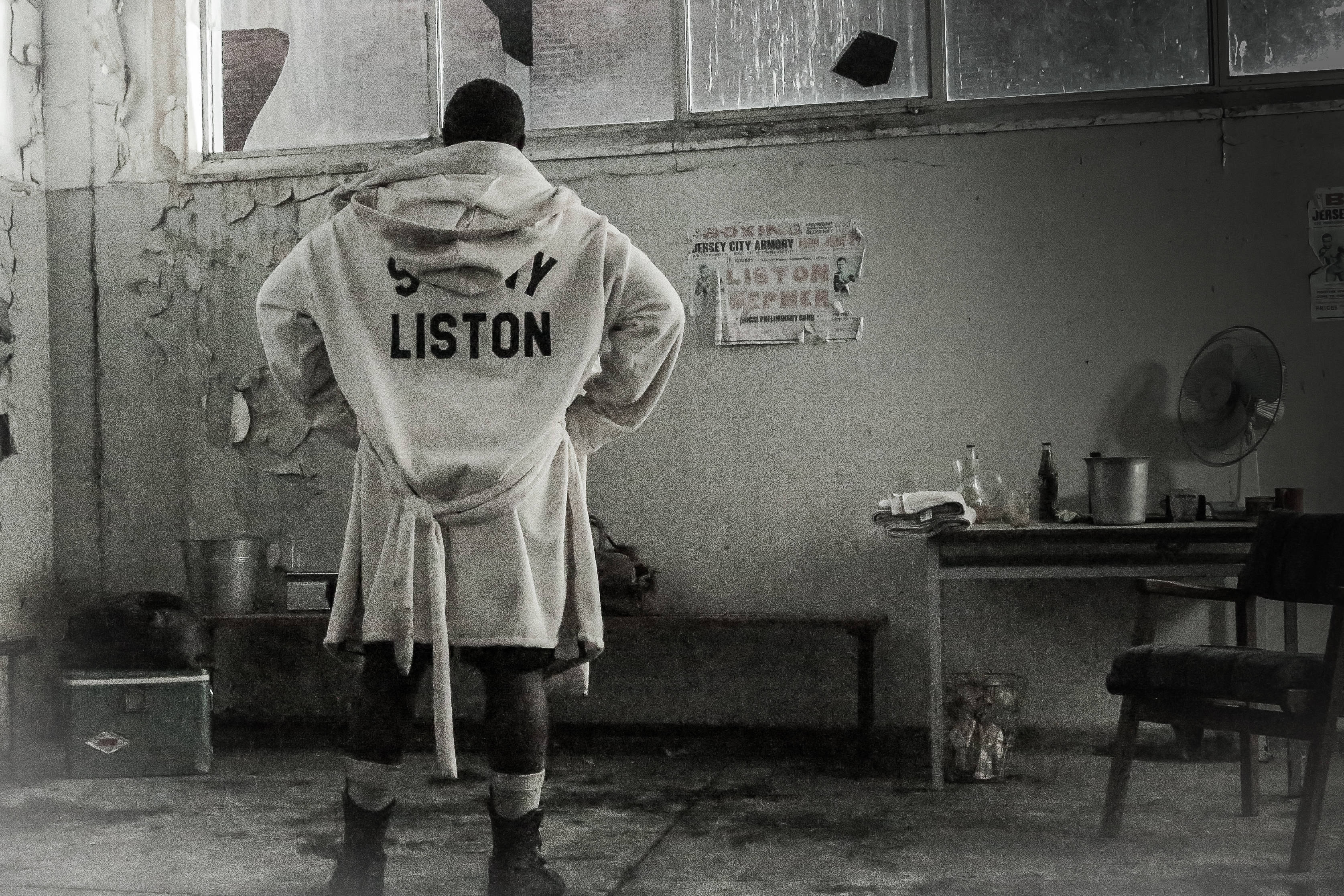 桑尼·利斯顿穿袍背影 壁紙 — 一位穿著 'SONNY LISTON' 長袍的拳擊手背對鏡頭，站在一個粗糙的黑白訓練室中。