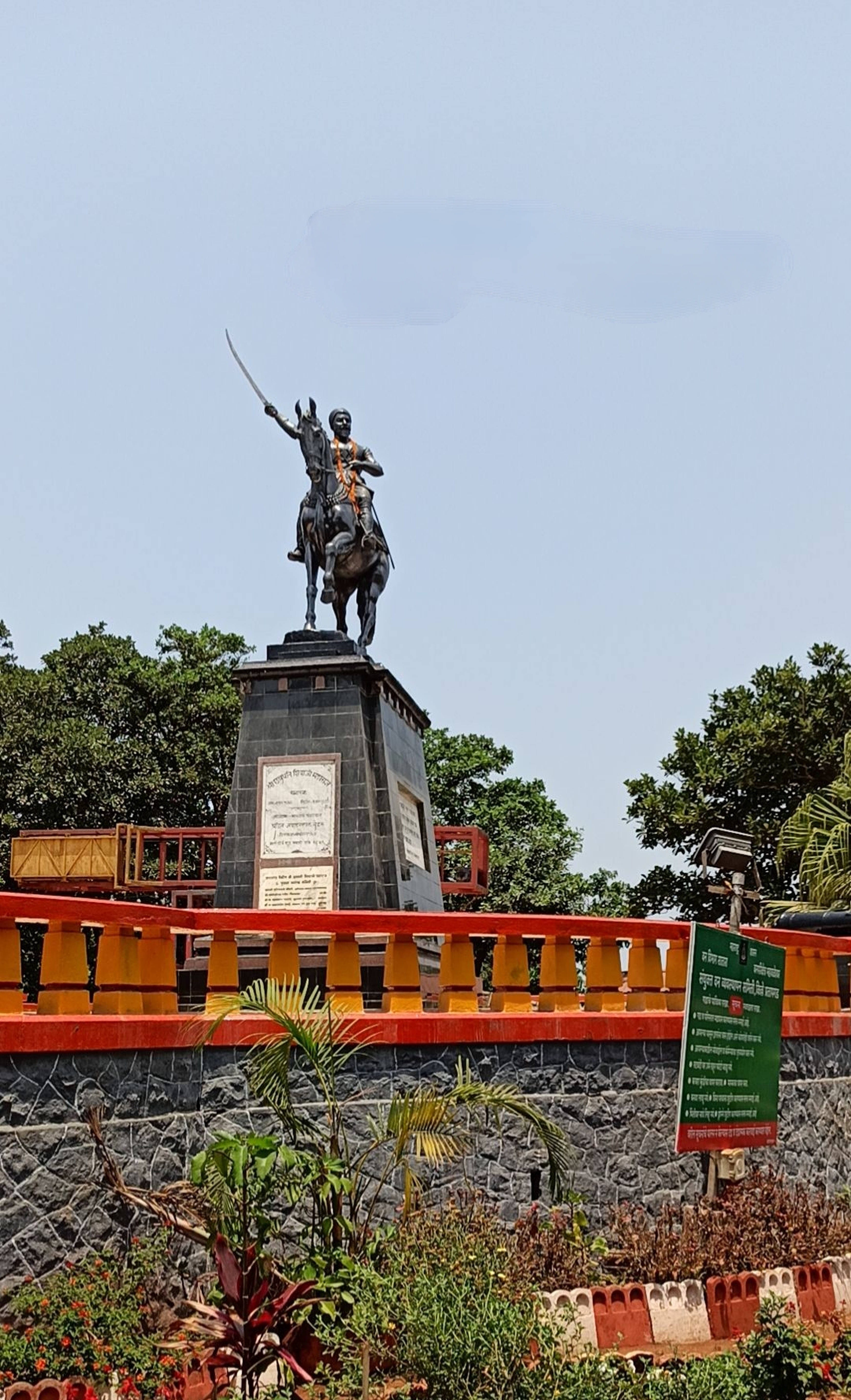 桑巴吉·馬哈拉吉之父騎馬雕像 壁紙 — 一座青銅騎馬戰士雕像，手持劍，矗立在石質基座上，周圍環繞著茂密的綠樹和色彩繽紛的欄杆，天空晴朗。