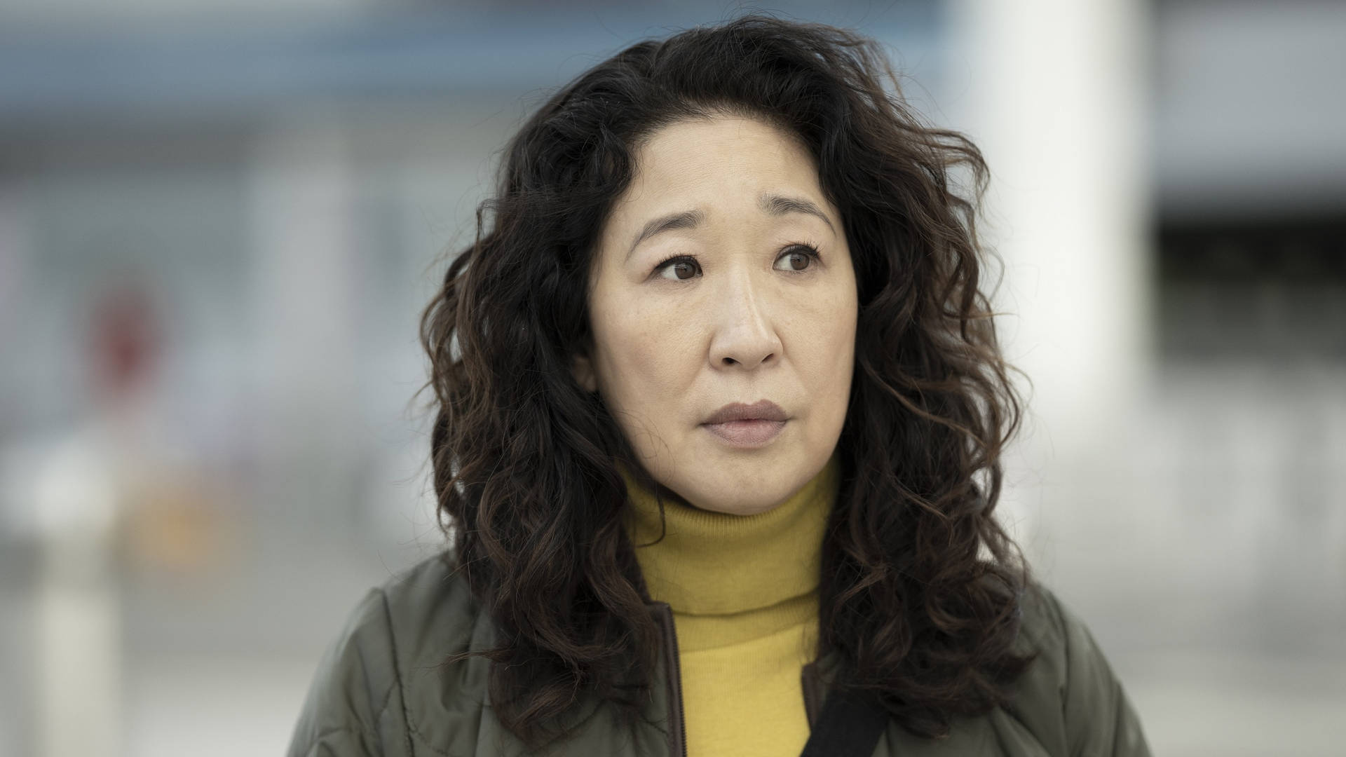 深色捲髮女性穿著黃色高領毛衣和外套，若有所思地望向遠方。