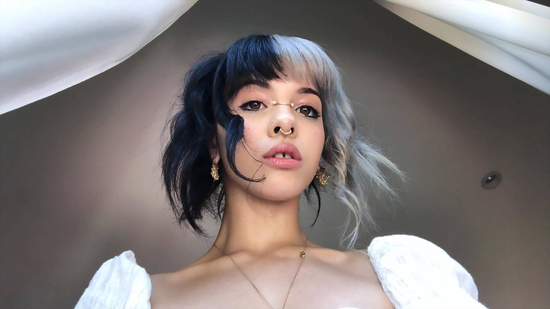 梅蘭妮·馬丁內斯鼻環特寫 壁紙 — 一位年輕女性，擁有引人注目的黑銀色頭髮和金色珠寶，在垂墜的白色布料下自信地擺姿勢。