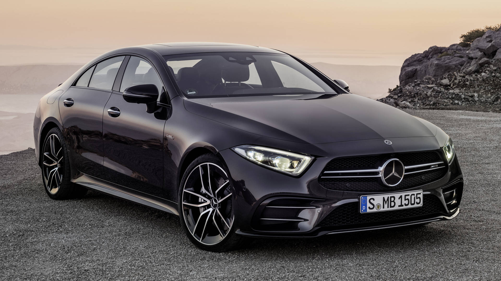 梅賽德斯-AMG CLS 53 壁紙 — 一輛時尚的黑色 Mercedes-AMG CLS450 轎車停在日落時分岩石懸崖旁的碎石路上。