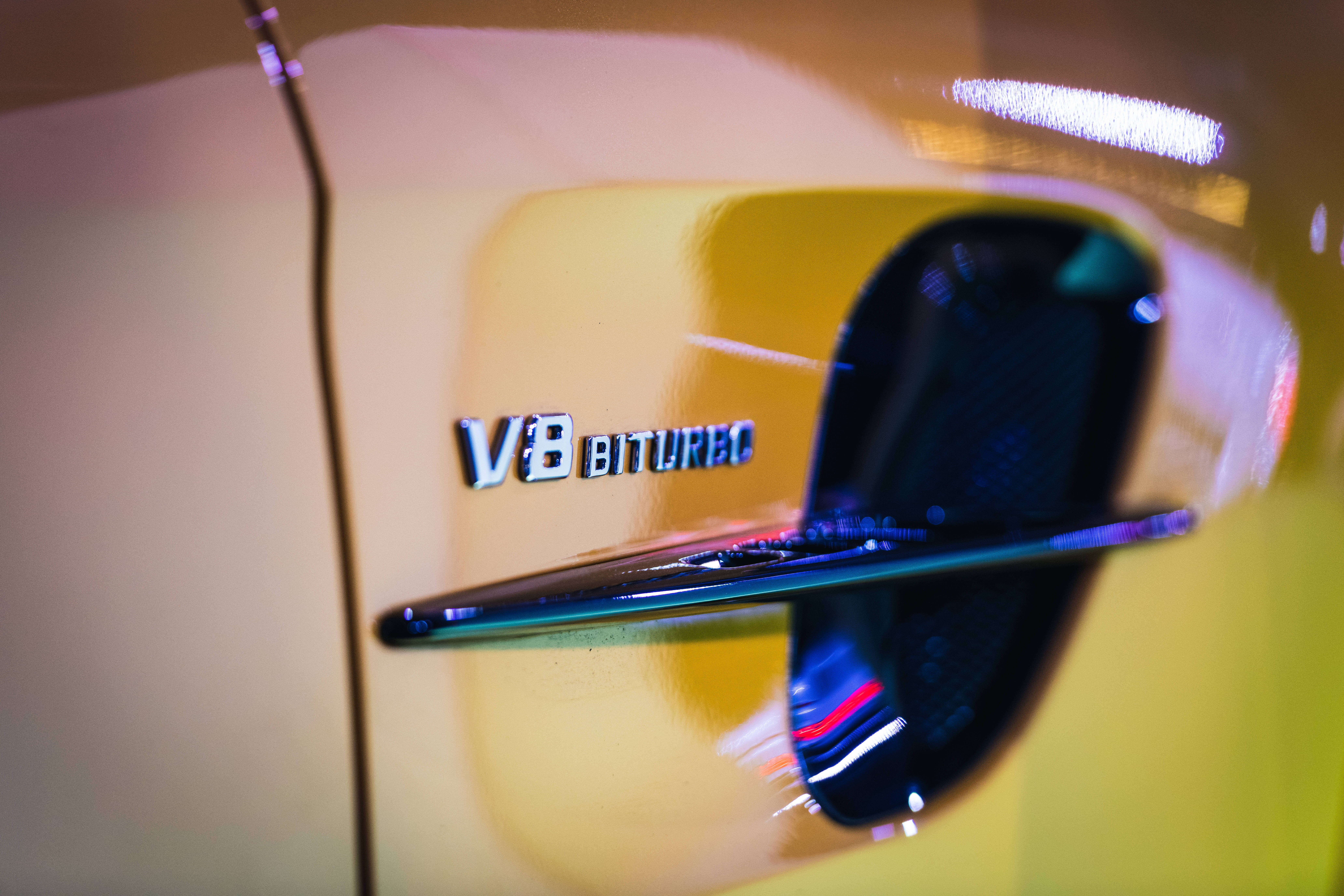 梅賽德斯-AMG V8渦輪增壓排氣管 壁紙 — 黃色車的V8 BITURBO徽章特寫，背景有模糊反射和霓虹燈。