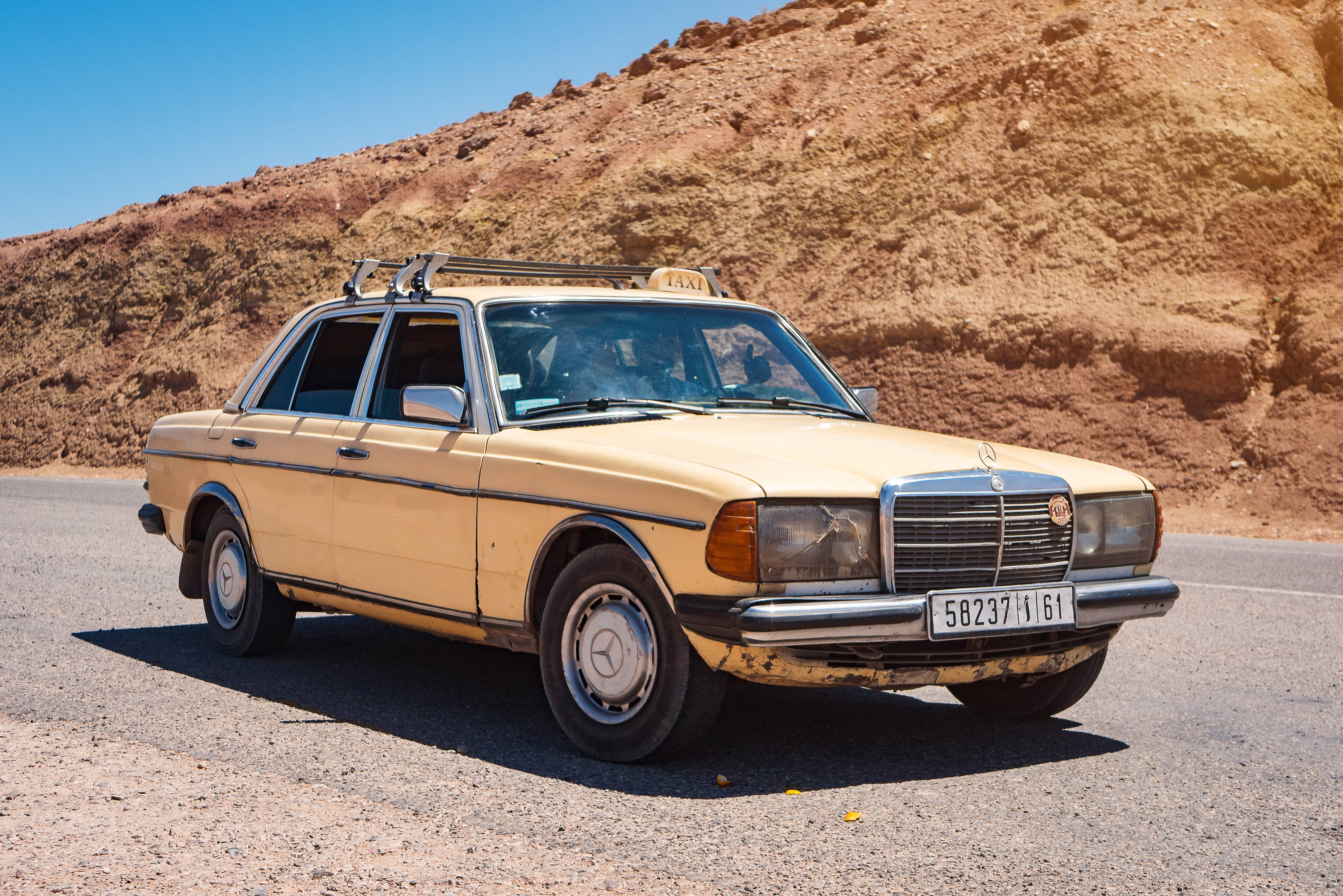 梅賽德斯-AMG W123 壁紙 — 一輛復古米色平治的士停泊在沙漠公路上，背景是晴朗的藍天和岩石山丘。
