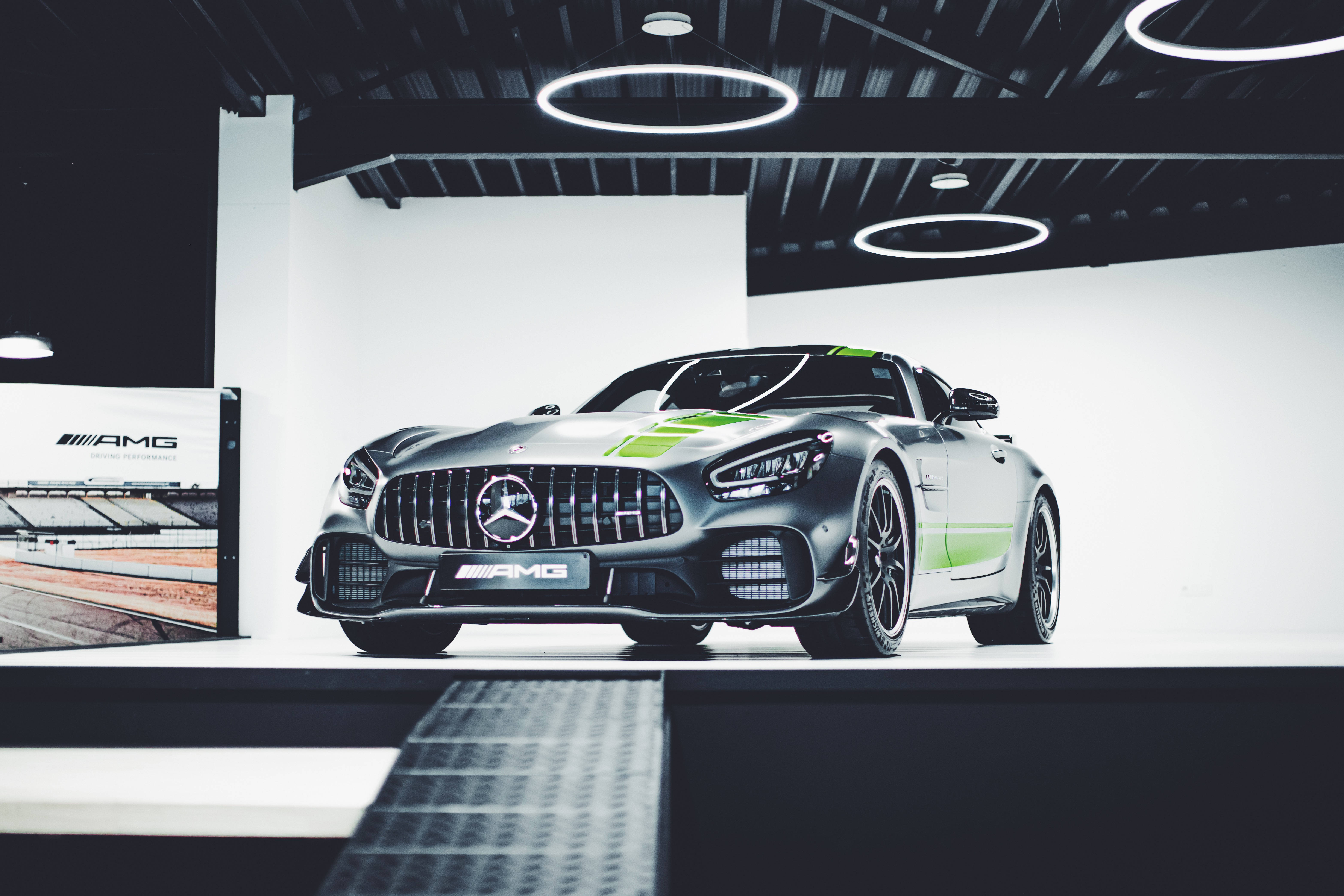 梅賽德斯-AMG 超跑 壁紙 — 一輛流線型銀色AMG GT賽車帶有綠色點綴，在圓形燈光下於極簡工作室環境中展示。