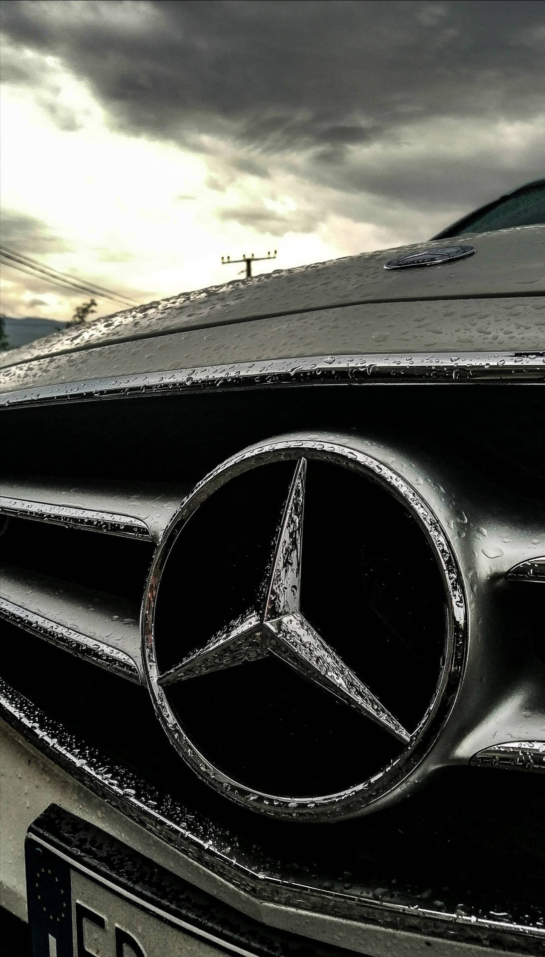 在暴風雨天空下，帶有雨滴的 Mercedes-Benz 汽車鬼面罩特寫。