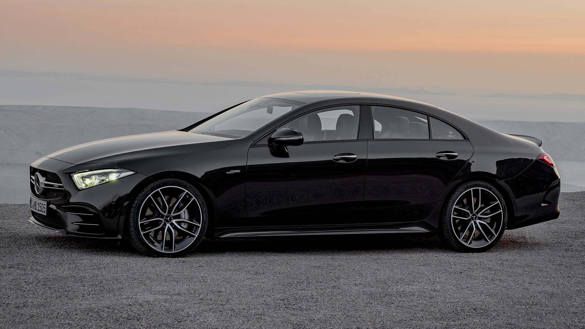 梅賽德斯-AMG 鋼灰色輪圈 壁紙 — 一輛時尚的黑色 Mercedes-AMG CLS 轎車在柔和的橙色日落天空下，在碎石觀景台上閃閃發光。