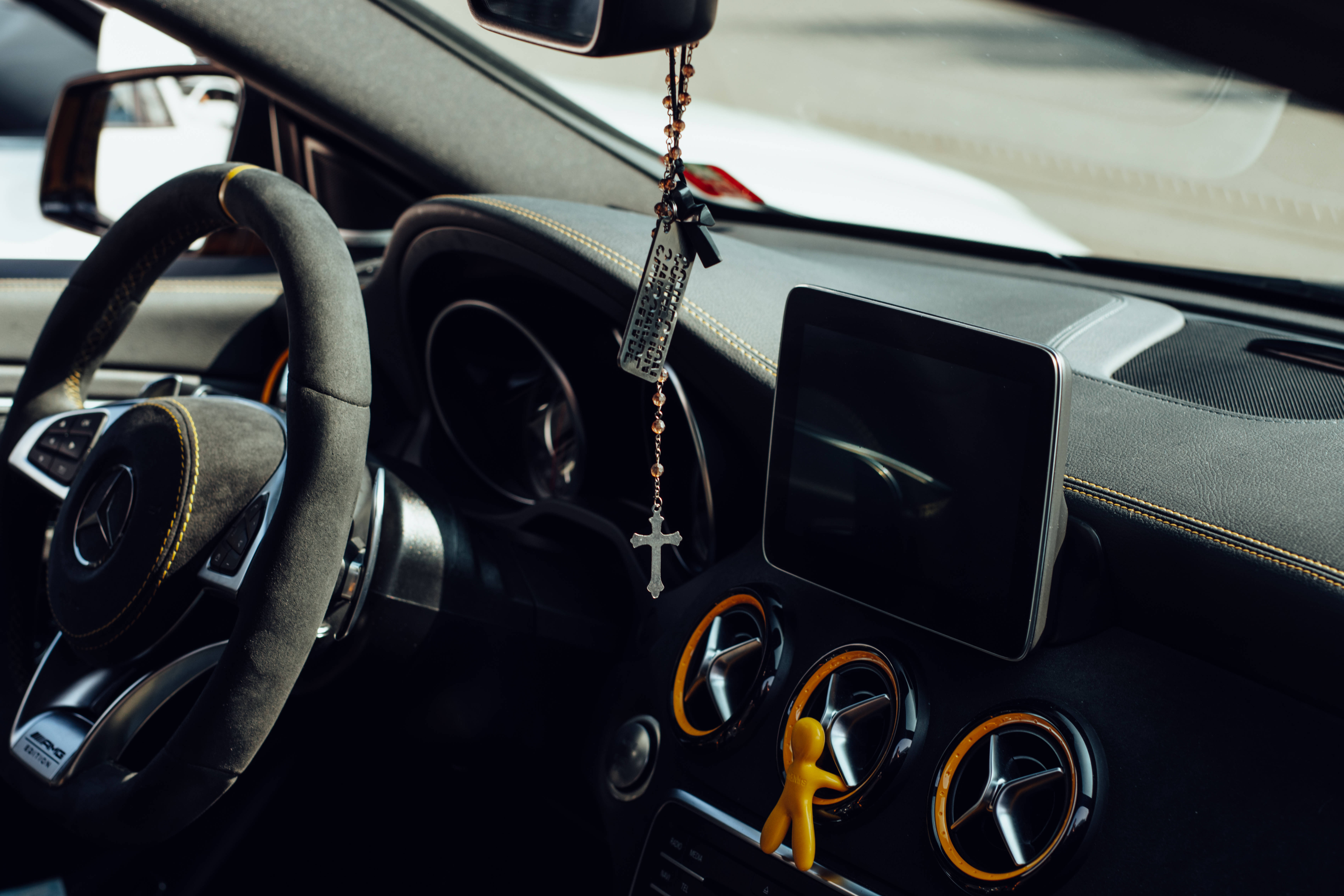 梅賽德斯-AMG 黑絨內飾儀表板 壁紙 — 豪華車內飾特寫，顯示方向盤、掛在後視鏡上的玫瑰經念珠和儀表板上的黃色公仔。