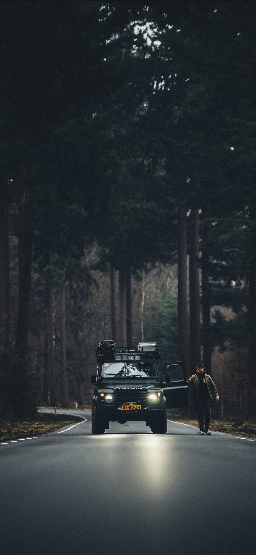 夜晚，一個人喺黑暗森林路上行喺一架停泊嘅綠色SUV旁邊。