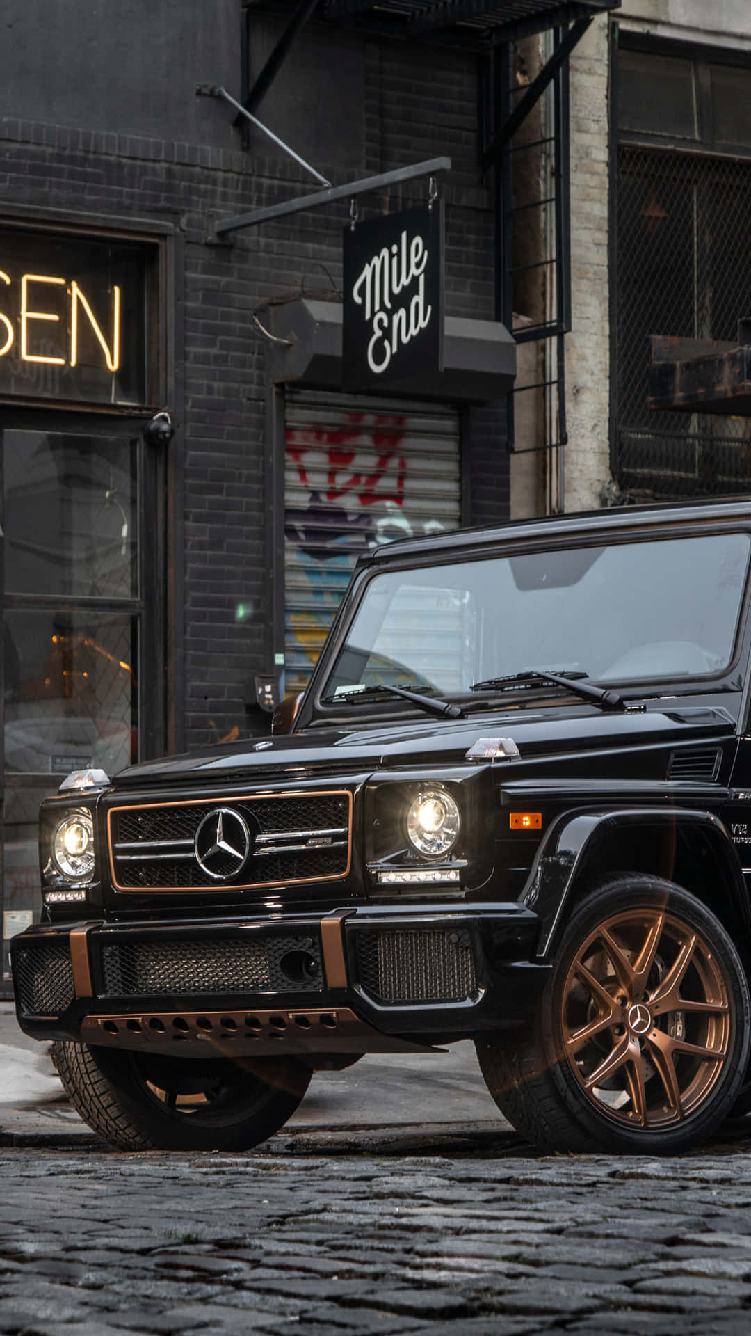 黑色Mercedes G-Wagen配金色輪圈夜間停泊在鵝卵石街道上。