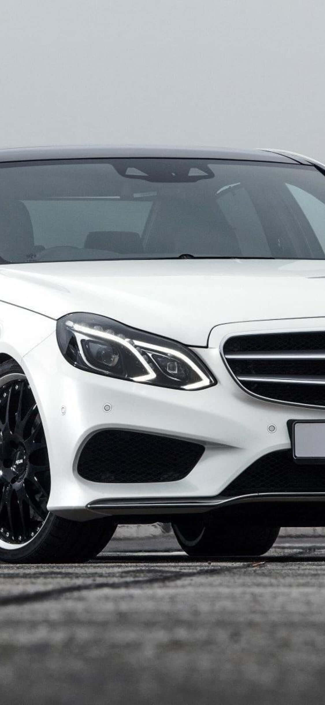 梅賽德斯-奔馳E級車前大燈 壁紙 — 一架時尚的白色 Mercedes-Benz 跑車配黑色輪圈，停泊在灰色瀝青路面上，天空多雲。