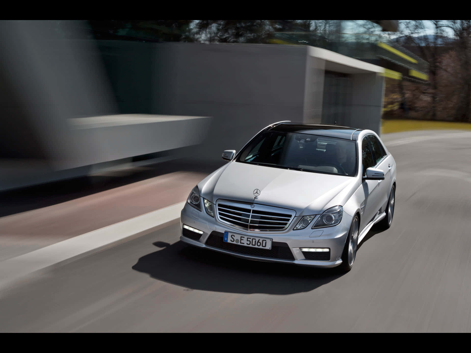 梅賽德斯-奔馳E級車穩穩駕馭 壁紙 — 一輛銀色 Mercedes E-Class 房車在現代化道路上疾馳，背景有模糊的動態和時尚建築。