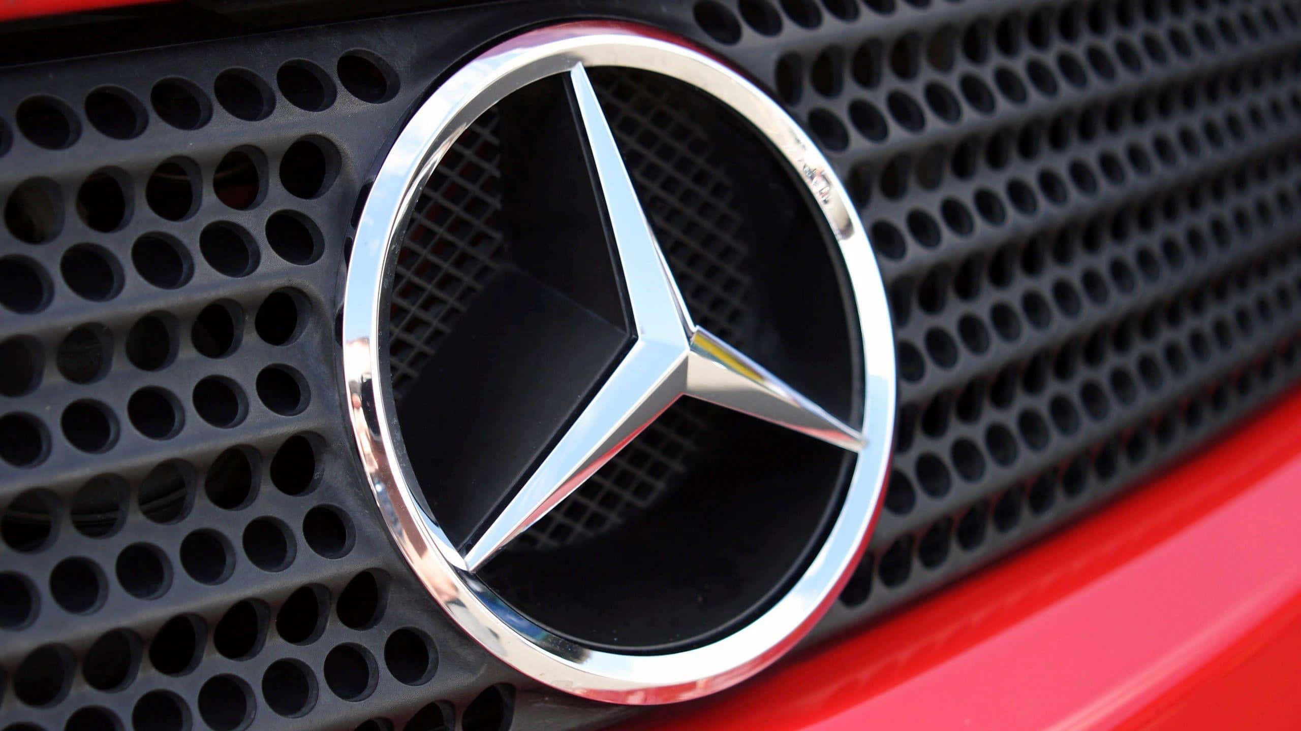 梅賽德斯-奔馳標誌 壁紙 — 特寫一個閃亮的鍍鉻 Mercedes-Benz 標誌，位於黑色穿孔格柵中央，下方可見紅色車身。