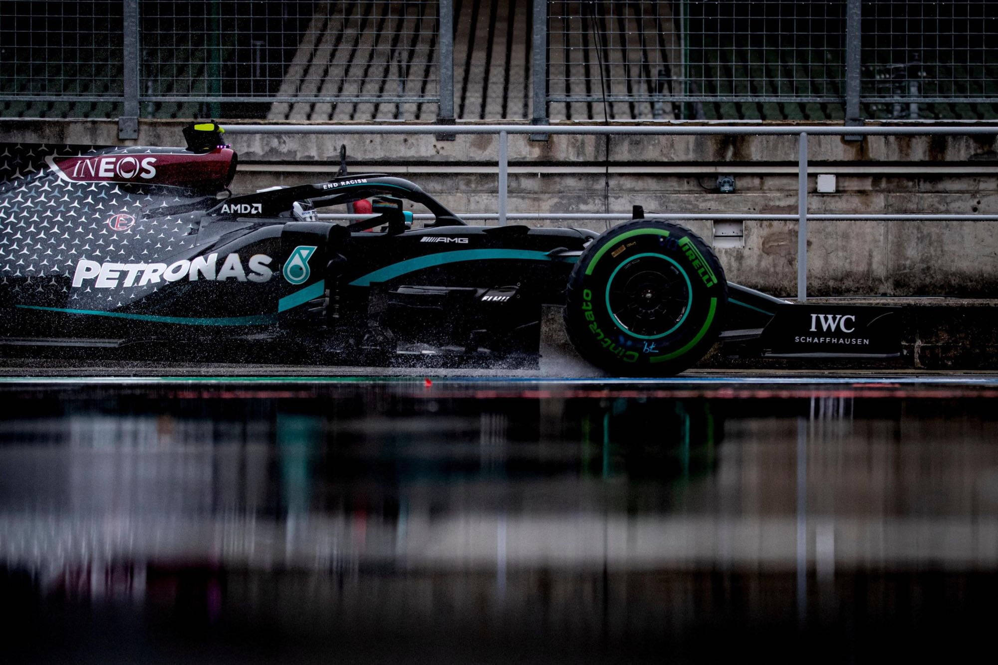 梅賽德斯AMG Petronas F1賽車賽道疾馳 壁紙 — 一架時尚的黑色和藍綠色一級方程式賽車在潮濕賽道上濺起積水，背景有混凝土護欄和圍欄。