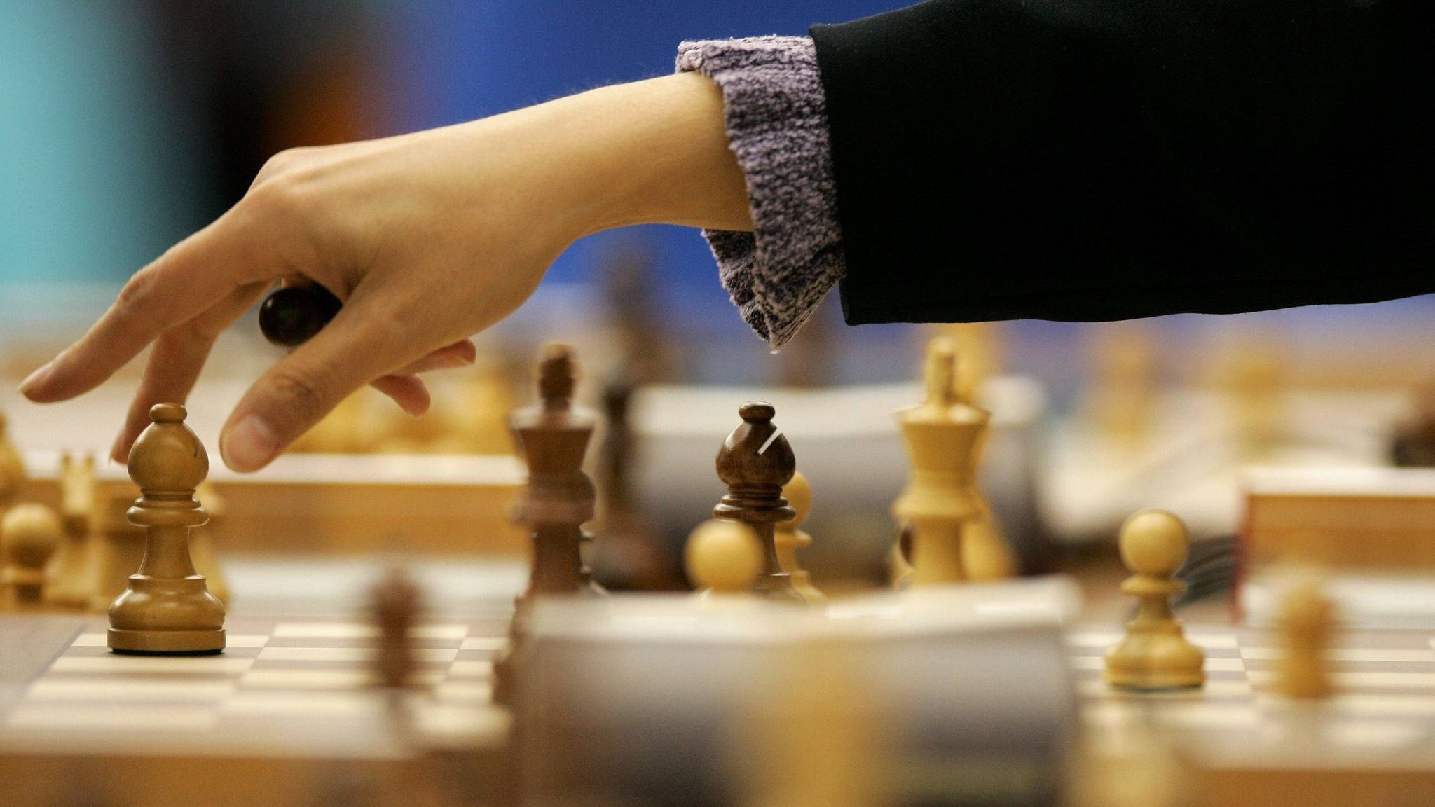 棋手的藝術：象棋的智慧 壁紙 — 一隻手伸向木製棋盤上移動國際象棋兵的特寫，背景有模糊的棋子。