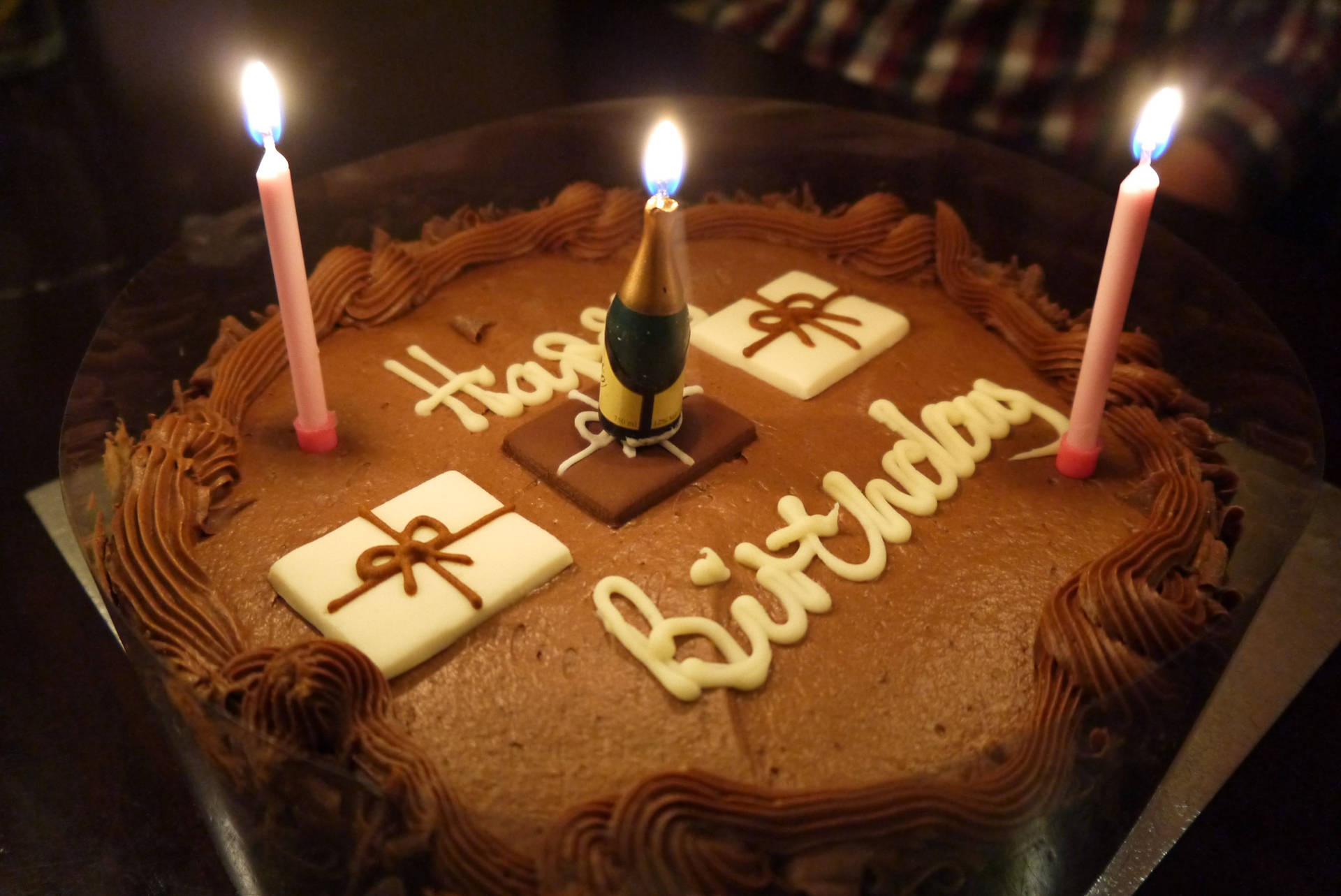 棕色大生日蛋糕與蠟燭 壁紙 — 在昏暗燈光下，一個帶有燃點蠟燭和「Happy Birthday」糖霜的朱古力生日蛋糕。