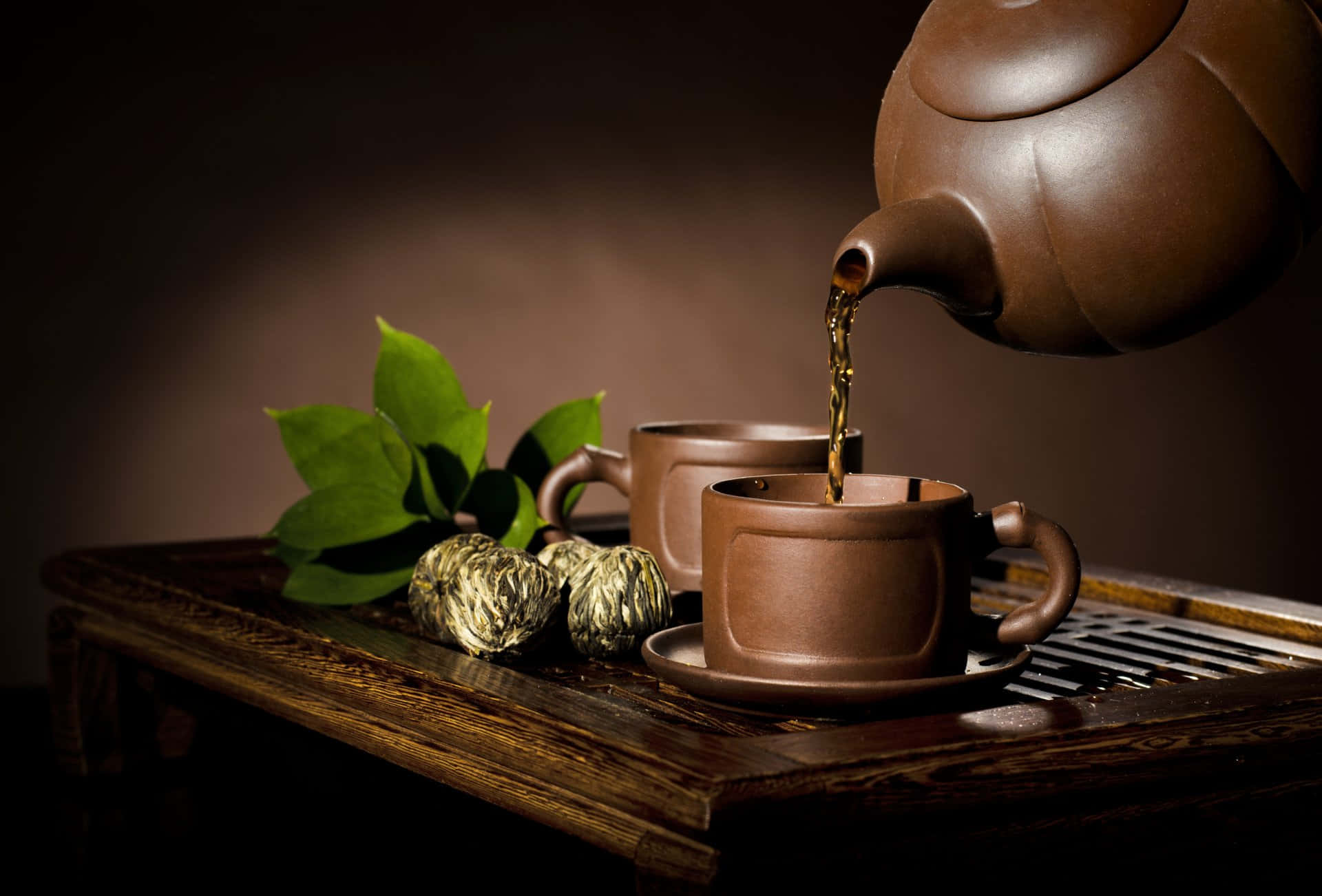 棕色茶杯與壺圖 壁紙 — 一個豐富的棕色陶壺將深色茶倒入木托盤上一個相配的杯子中，旁邊有綠葉和壓縮茶球。