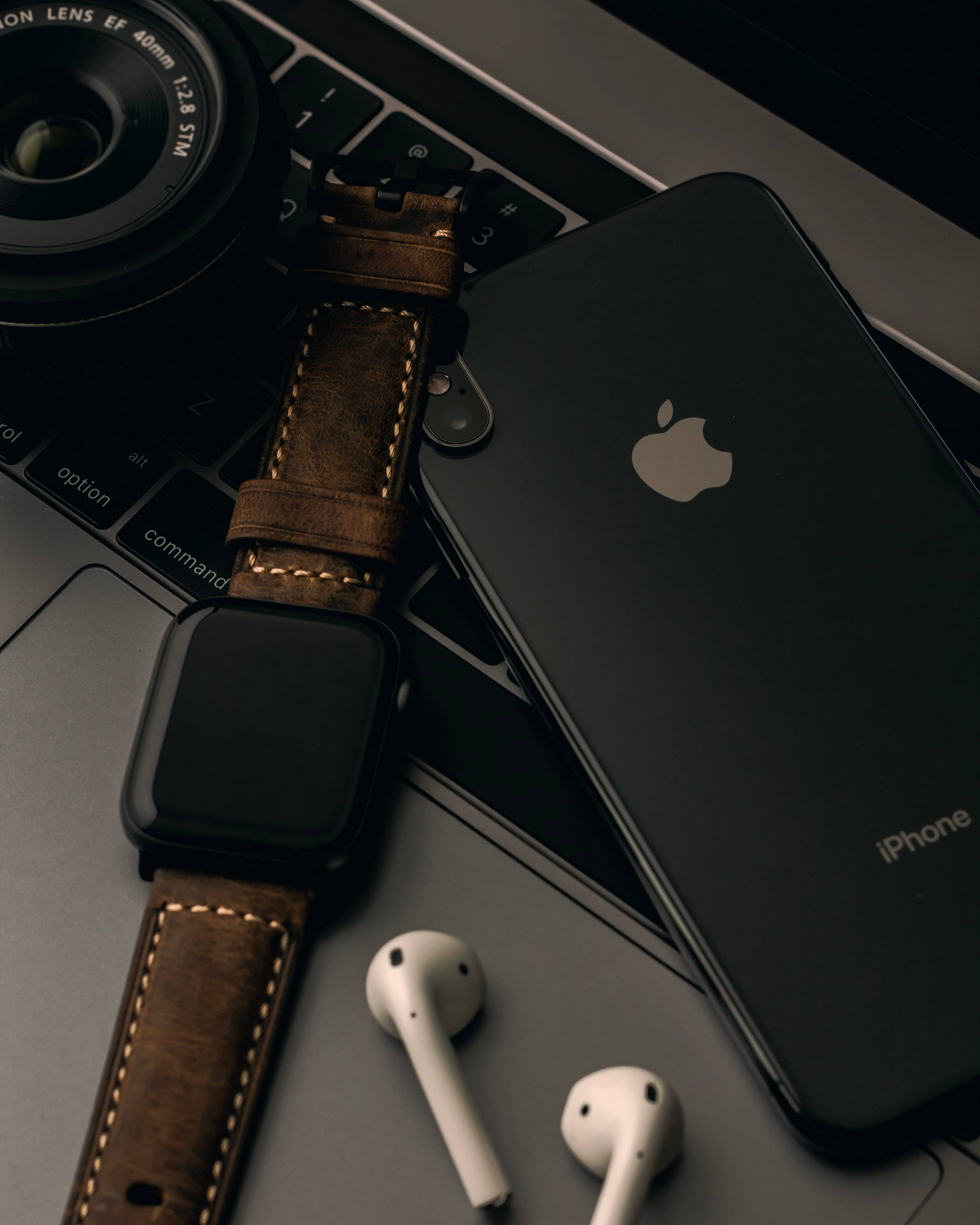 棕色錶帶 Apple Watch 4K iPhone 11 壁紙 — 一部時尚的黑色iPhone和棕色皮革錶帶的智能手錶放在筆記型電腦鍵盤上，旁邊有白色耳機和相機鏡頭。