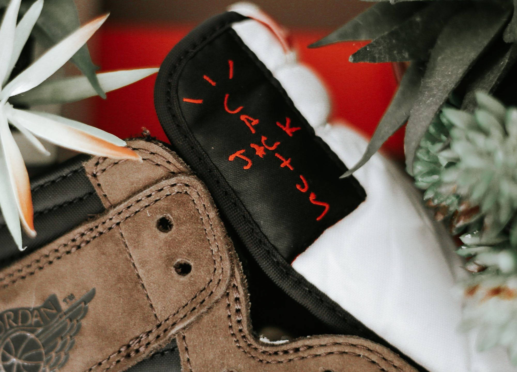 Cactus Jack Air Jordan 壁紙 — 棕色麂皮Jordan運動鞋近距離特寫，黑色標籤上有手寫橙色文字，藏在白色和綠色葉子之間。