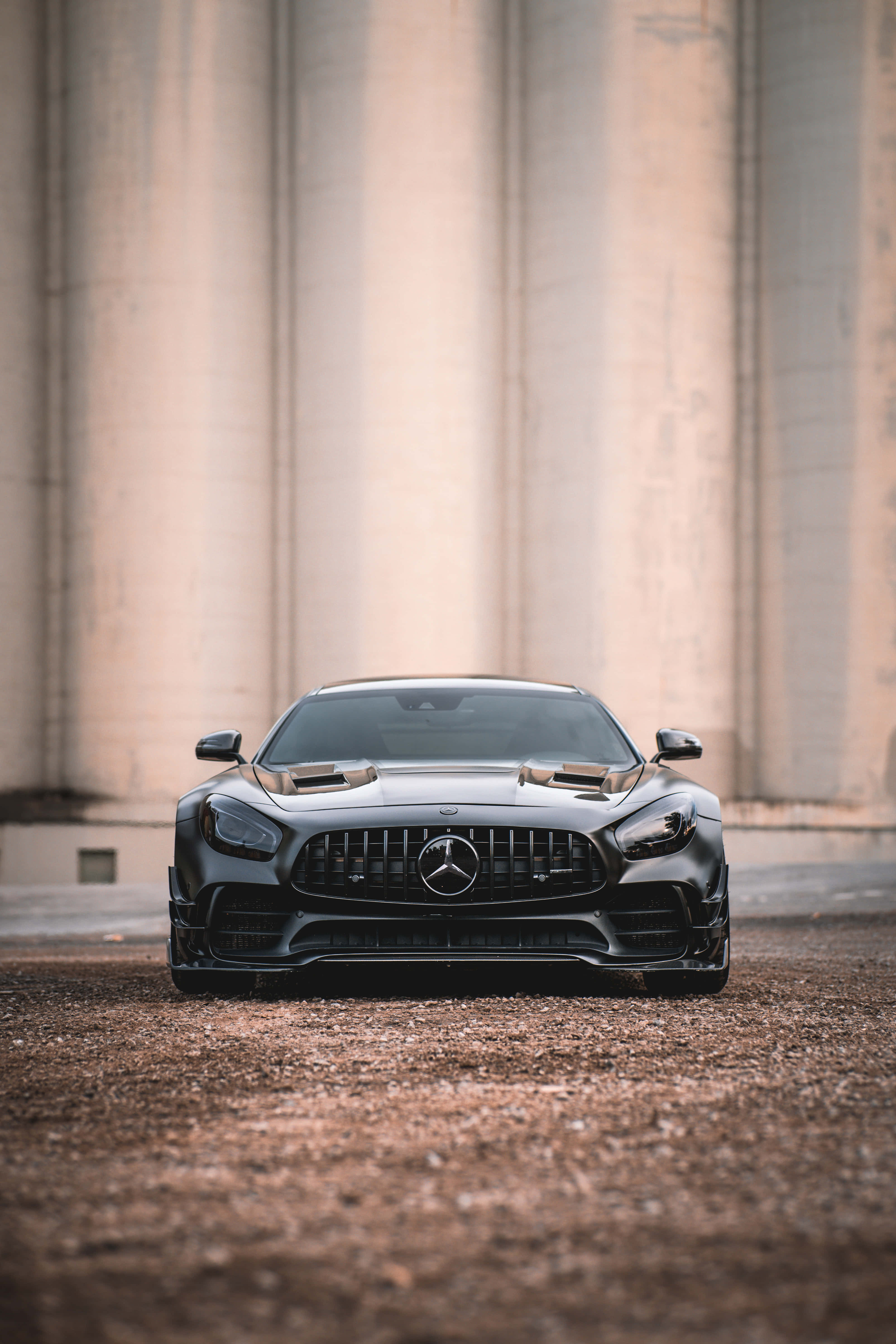 棕調黑AMG GT-R安卓背景 壁紙 — 一輛時尚的黑色Mercedes-AMG GT R跑車在碎石上朝前，位於高聳的混凝土柱下方。