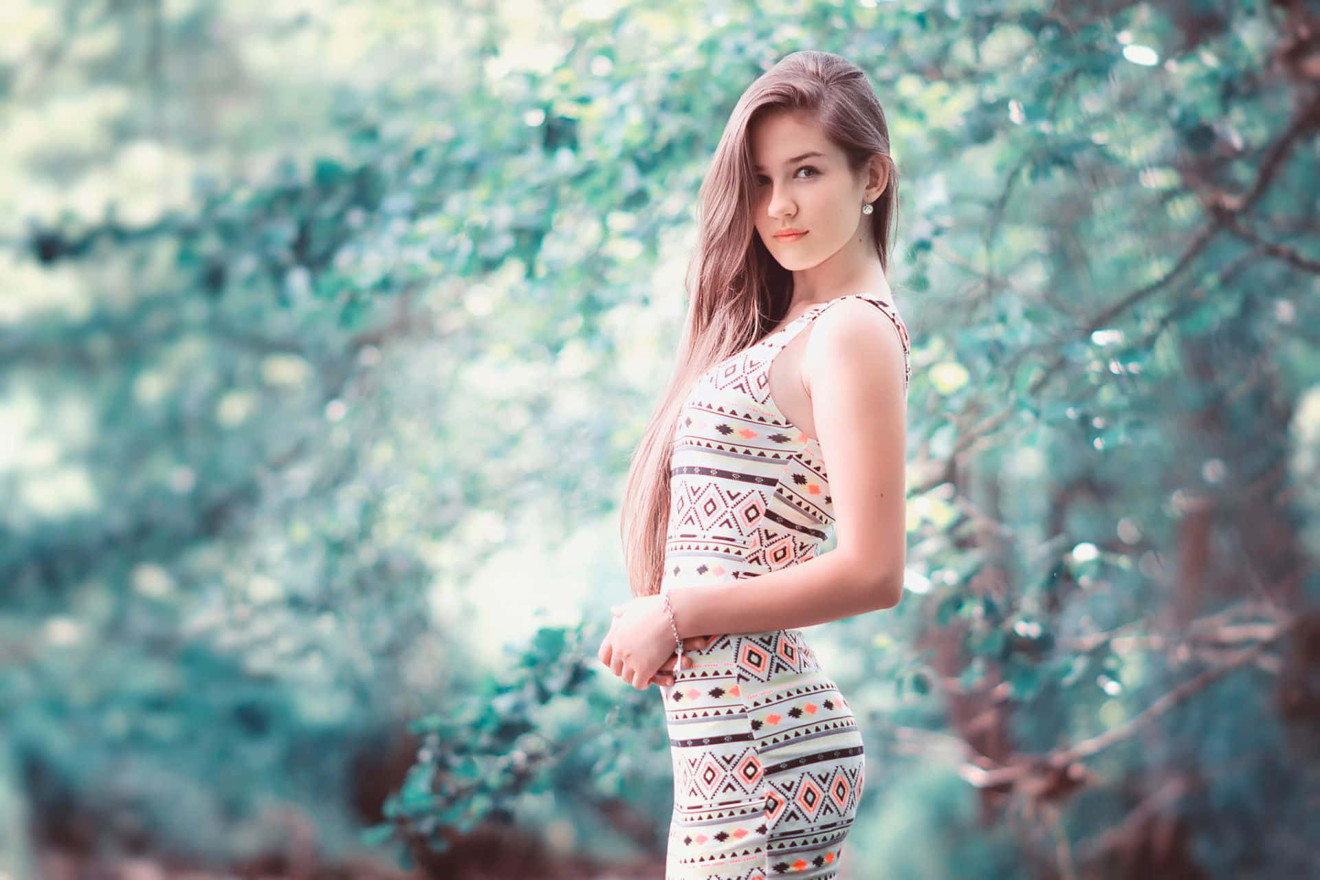 森林中的少女照片 壁紙 — 一位身穿圖案連衣裙的年輕女子優雅地站在陽光斑駁的樹木間，身後是柔和的散景樹葉。