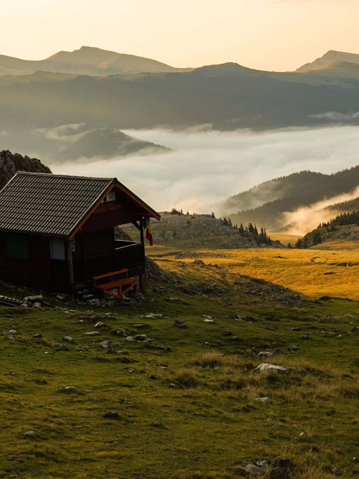 森林中的自然美景 壁紙 — 一座木造山間小屋棲息在雲霧之海上，金色陽光照耀著起伏的丘陵和遠方的山峰。