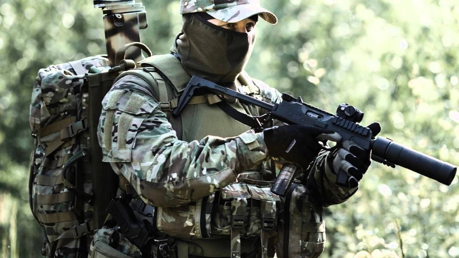 森林中的軍隊 壁紙 — 一名身穿全套迷彩裝備的士兵，透過茂密的綠色樹葉瞄準一支帶有消音器的步槍。