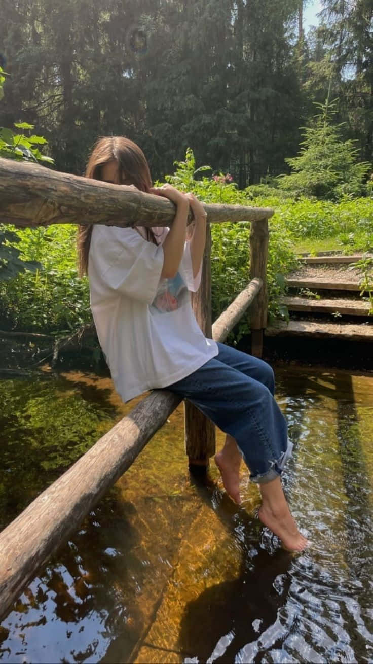 年輕女性赤腳坐在森林中小溪上的木橋欄杆上。