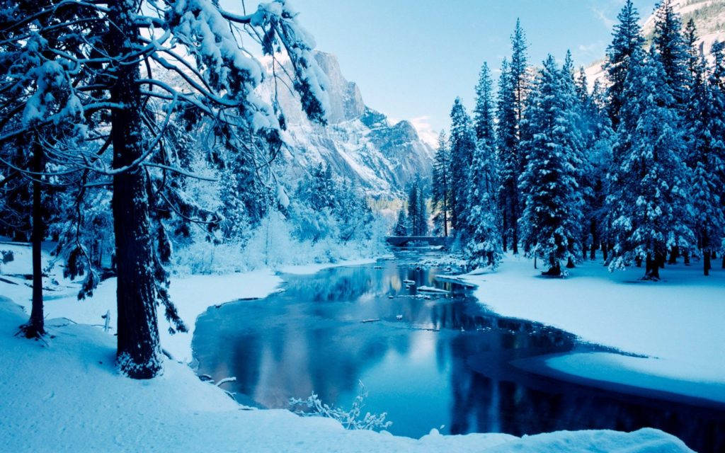 森林溪流冬日桌面壁紙 壁紙 — 寧靜的冬日景象，白雪覆蓋的松樹排列在平靜的河流兩旁，河流映照著冰藍色的山脈，天空清澈。